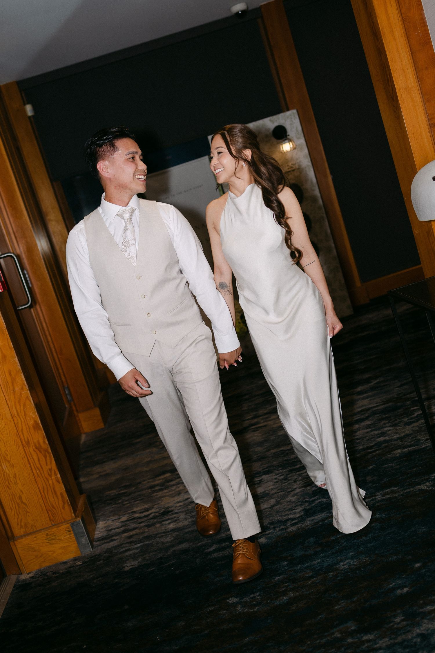 Couple in elegant wedding attire holding hands and smiling while walking through a venue hallway.