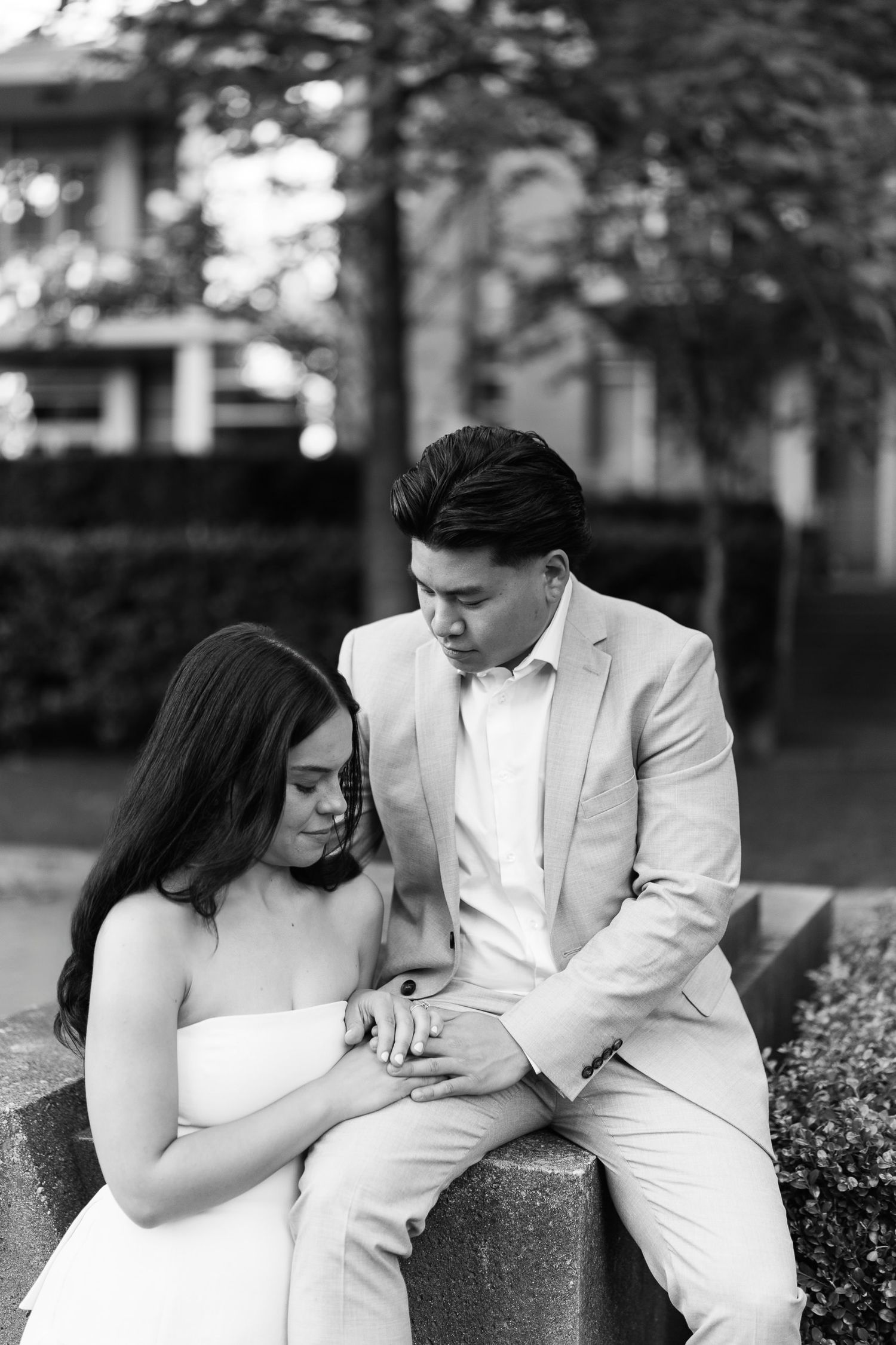 Black and white engagement portrait of a couple sitting together on a stone ledge outdoors, holding hands and looking down.