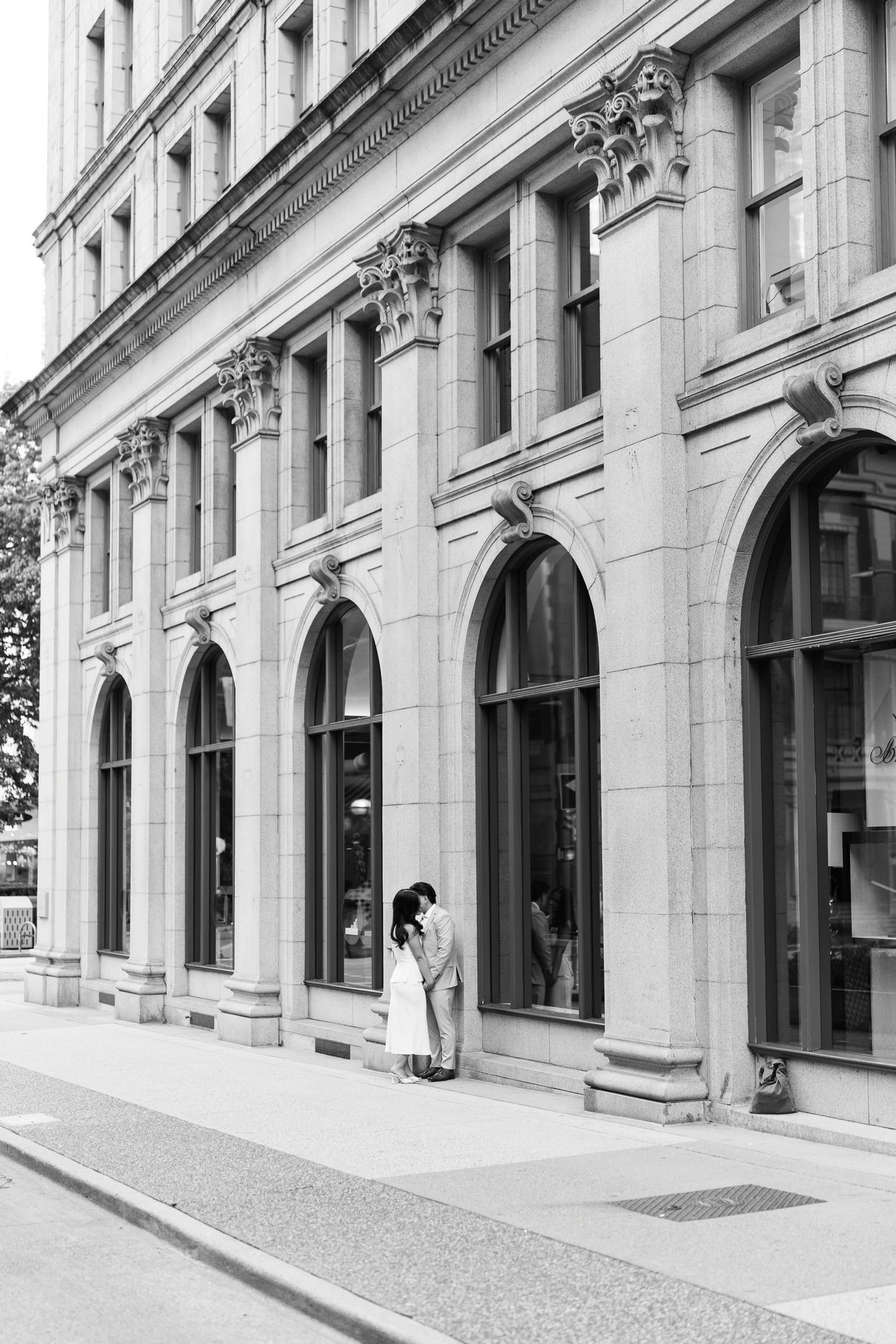 Black and white photo of a couple embracing beside a grand stone building with tall arched windows on an urban street.