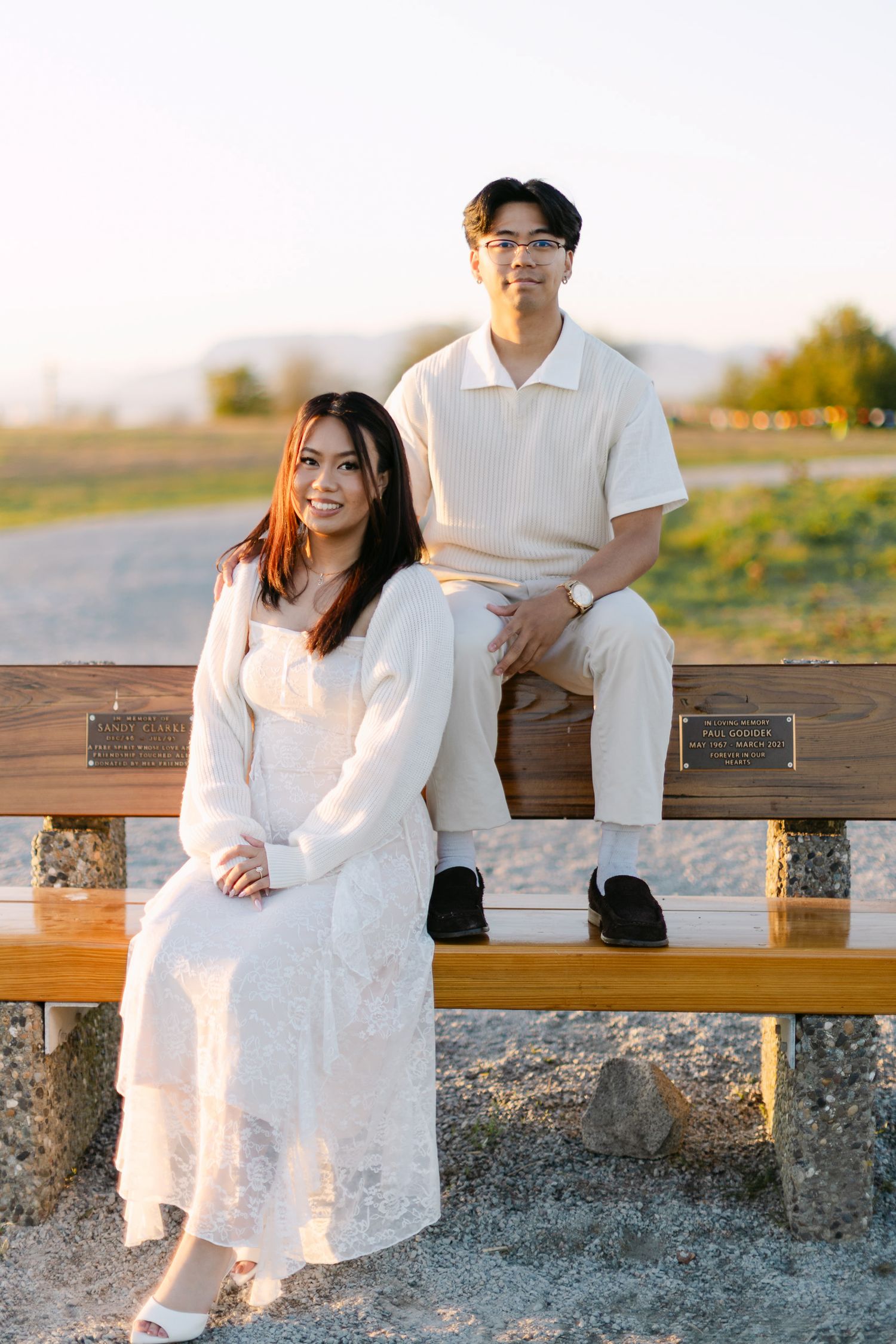 A smiling woman in a white lace dress sits on a wooden bench while a man in cream sits on the backrest behind her at sunset.