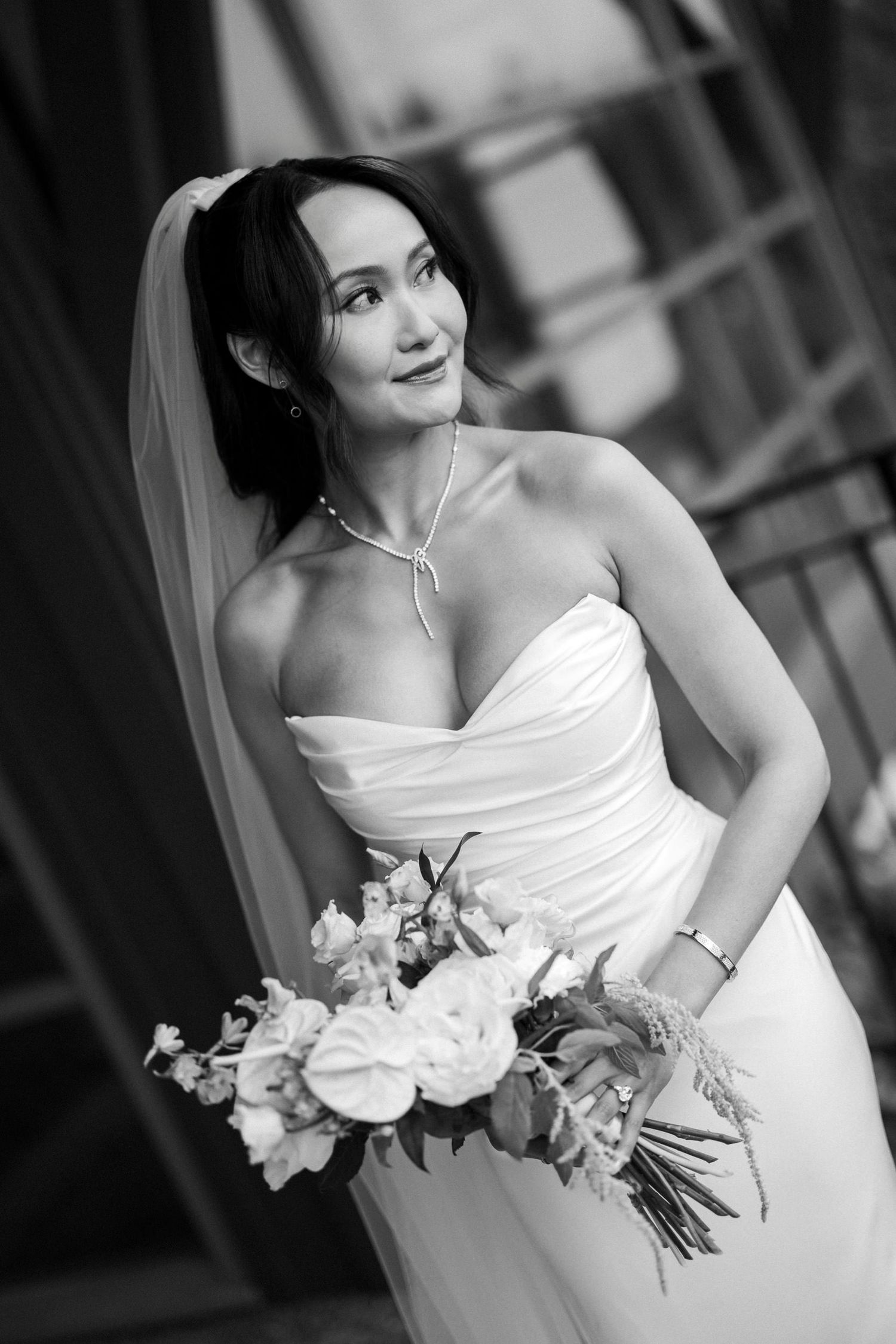 Elegant bride in strapless white wedding gown holding bouquet, wearing veil and necklace, black and white portrait photo.
