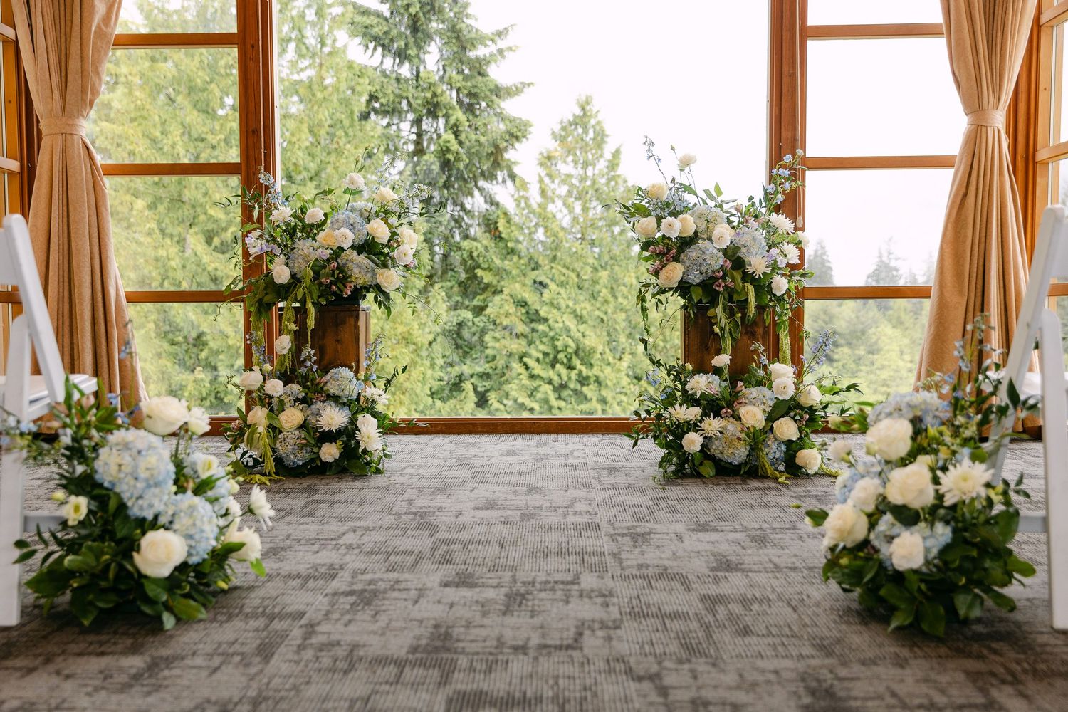 Elegant wedding ceremony setup with white floral arrangements, greenery, and large windows overlooking lush trees.