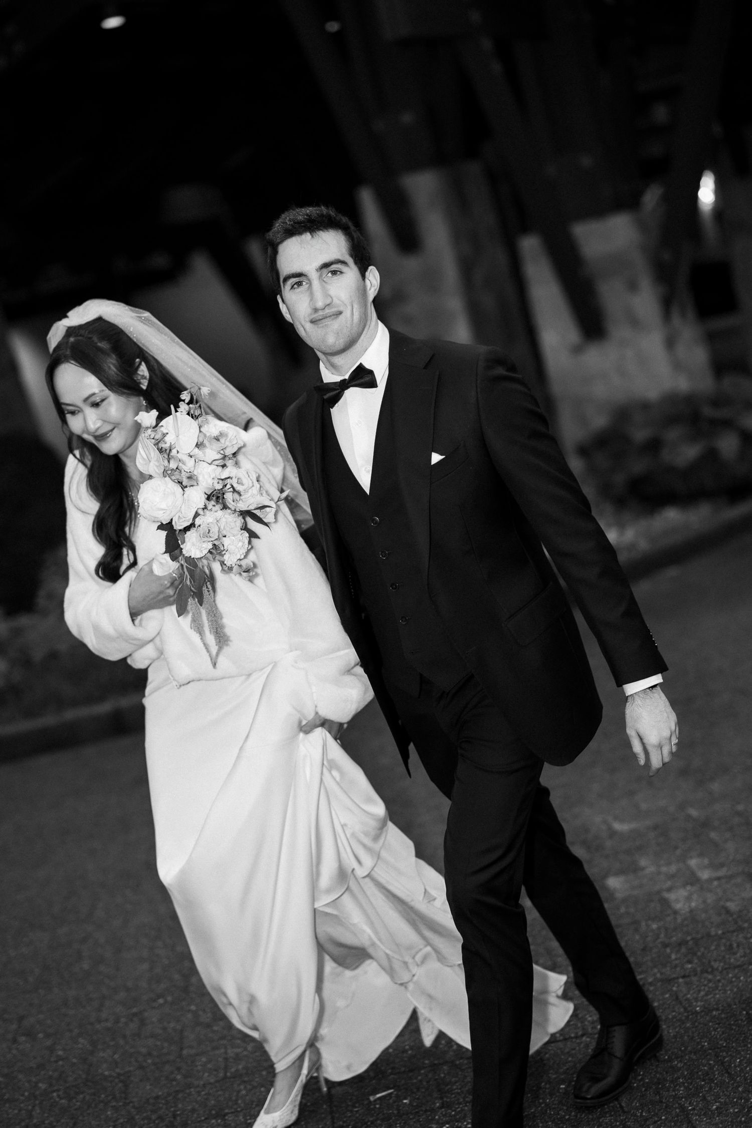 Black and white photo of a bride and groom walking together, she holds a bouquet, he wears a tuxedo.