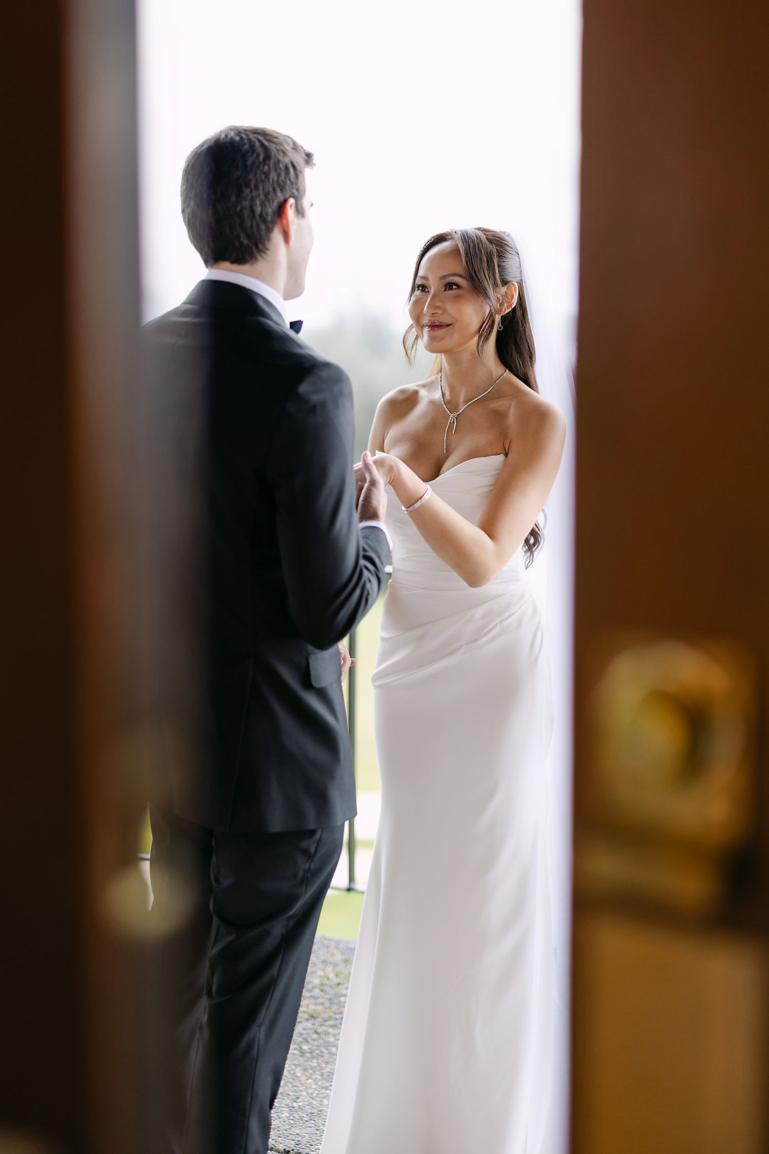 Bride in elegant white strapless wedding gown smiling at groom through a doorway.