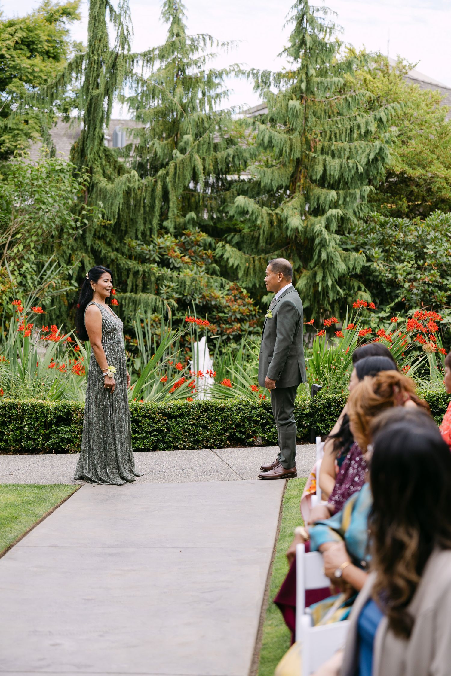 Outdoor wedding ceremony in a lush garden with couple facing each other on a path, guests seated nearby.