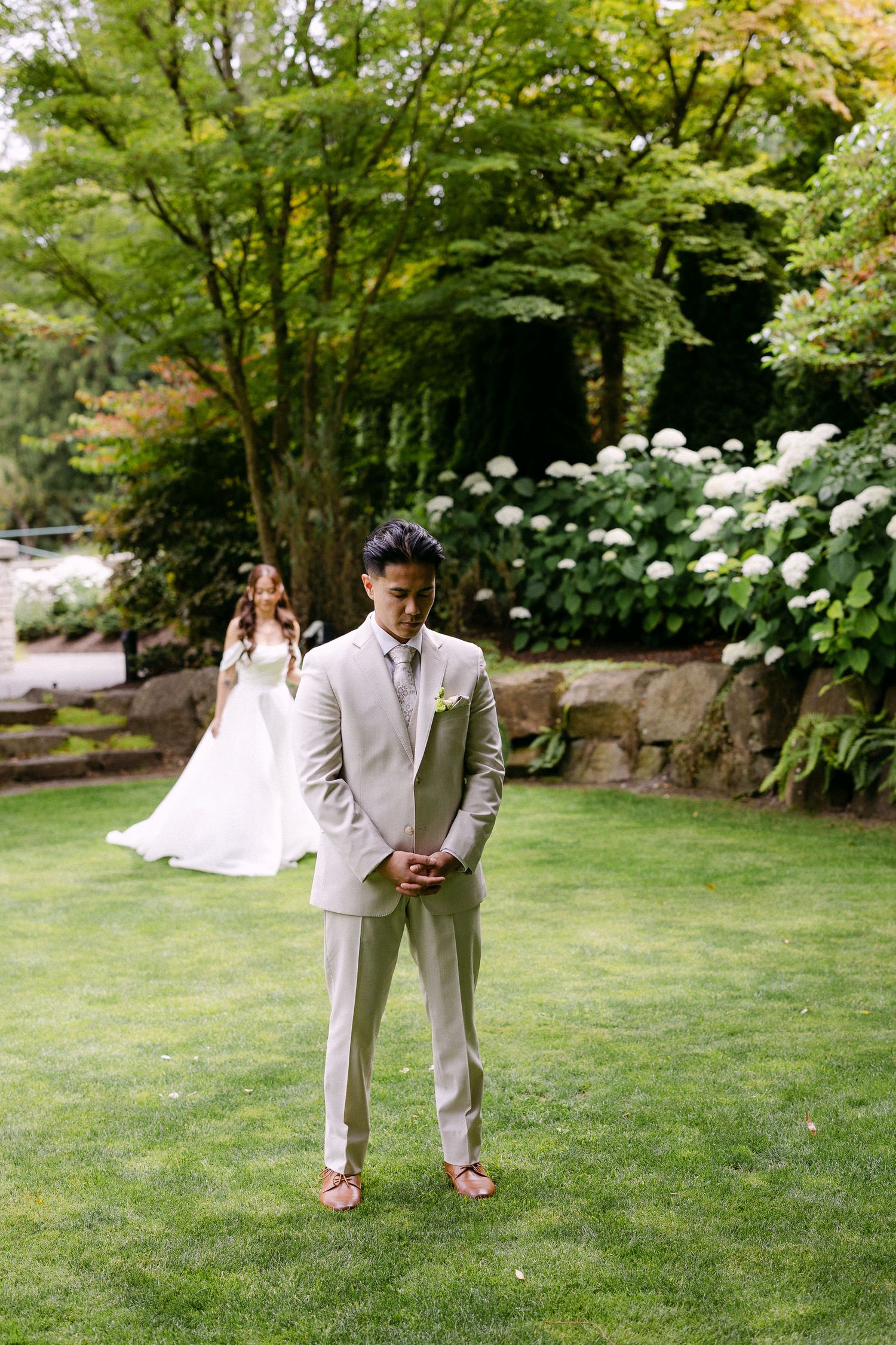 Groom in beige suit stands on green lawn as bride in white dress approaches during wedding first look.