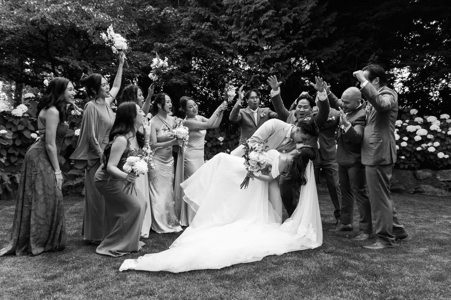 Wedding party celebrates as groom dips bride surrounded by cheering bridesmaids and groomsmen outdoors.