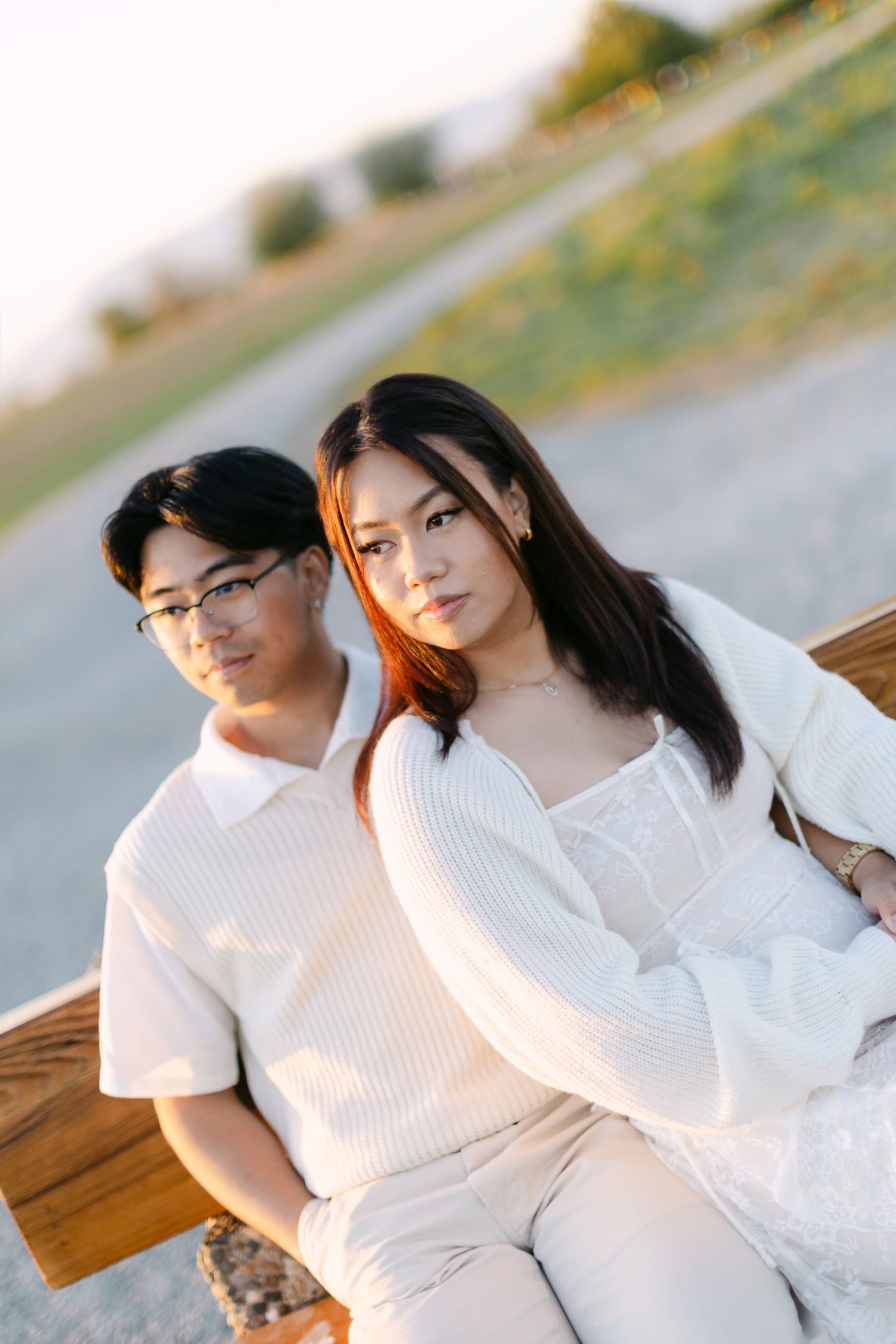 Couple sits close together on a wooden bench outdoors in soft golden light, dressed in matching cream tones.