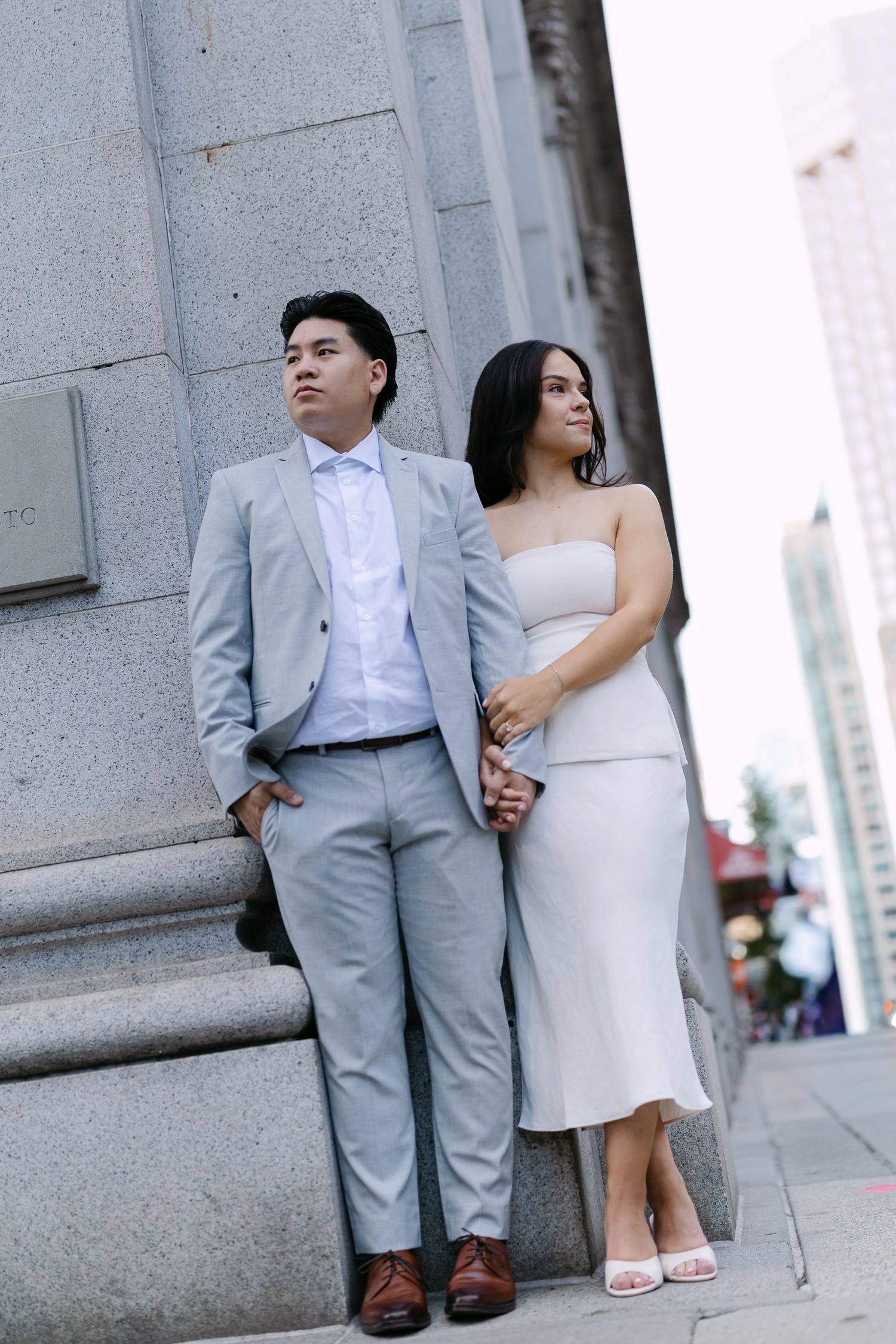 Engagement portrait of a couple holding hands beside a granite building column, with city skyscrapers in the background.