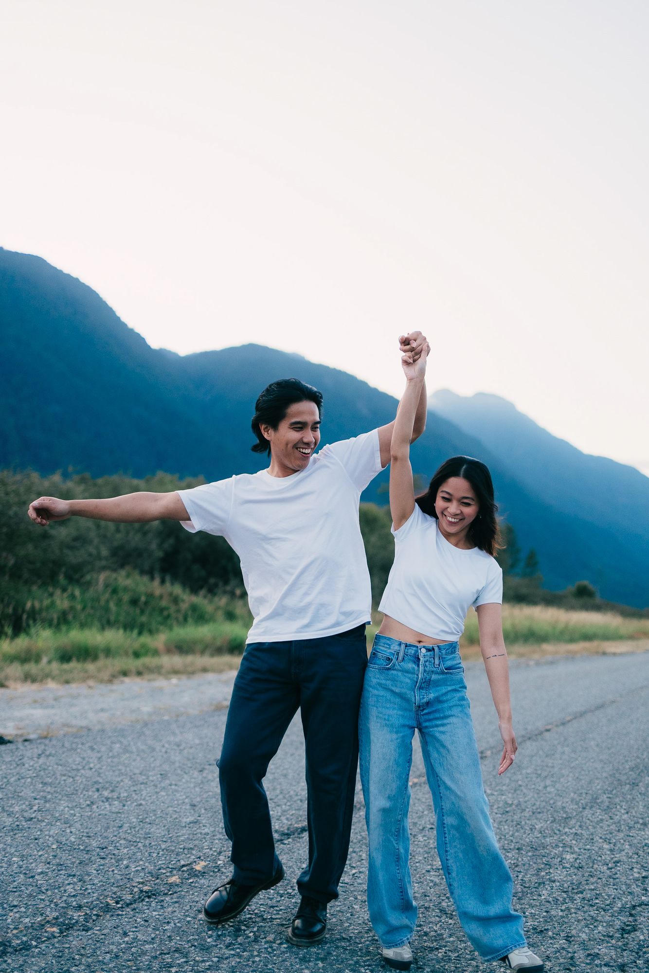 Smiling couple in white t-shirts and jeans hold hands raised high while dancing on a quiet paved road with mountains behind them.