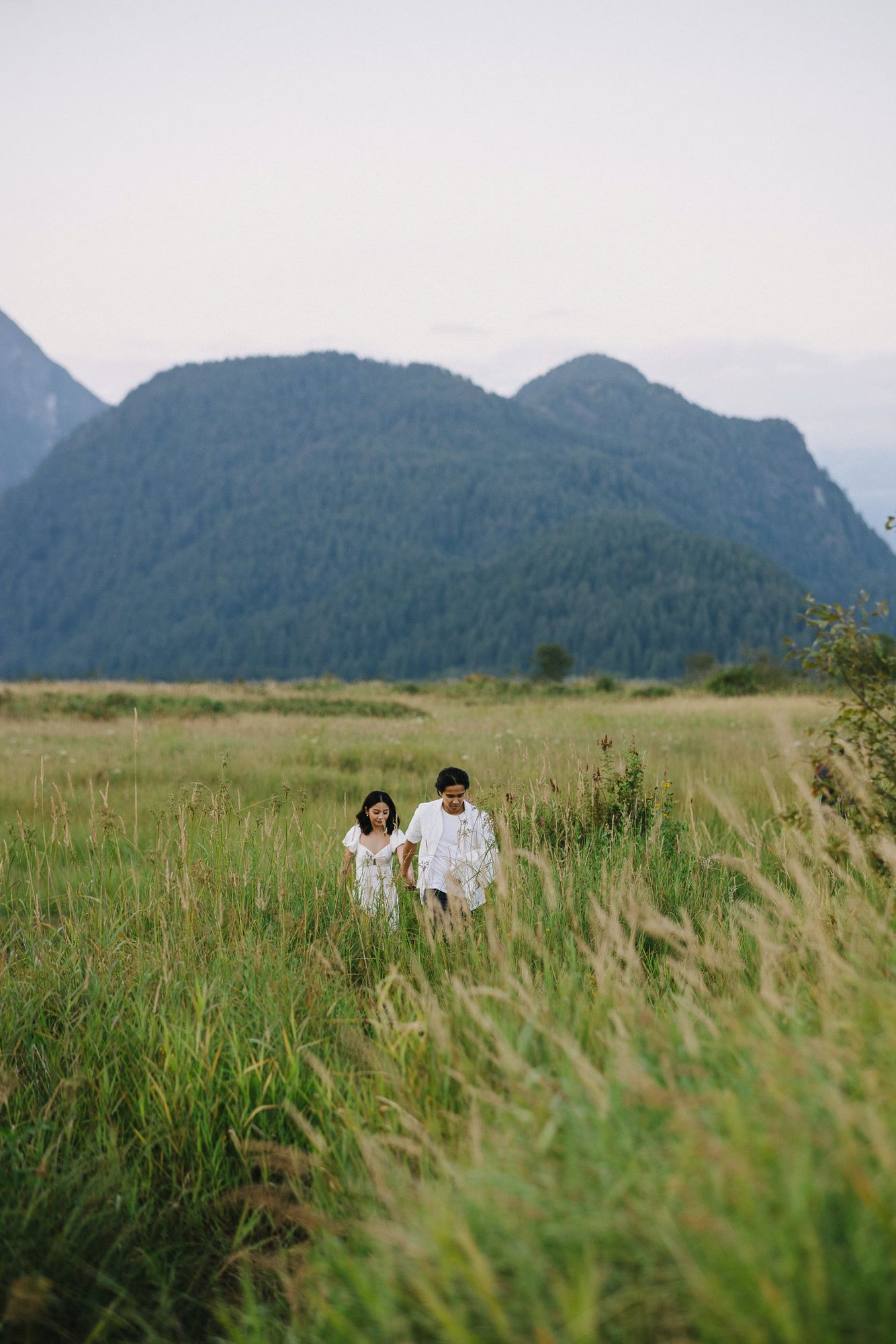 Couple in white walking through tall green grass in a wide valley with forested mountains rising in the background.