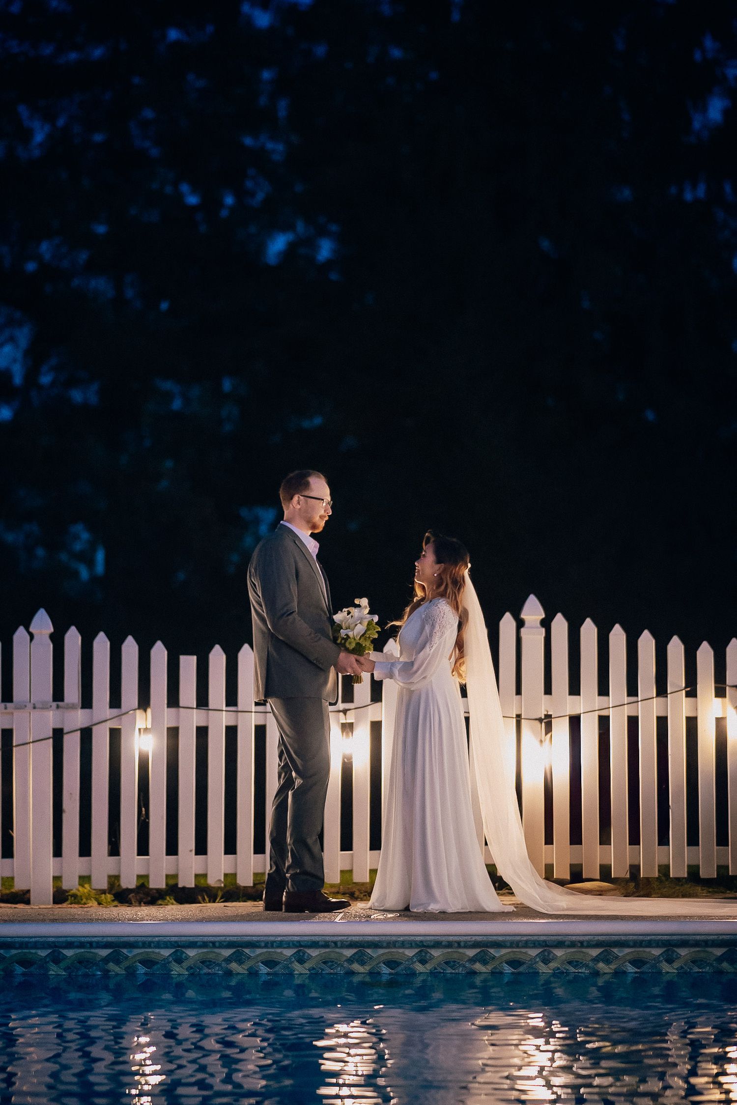 A romantic evening wedding ceremony takes place beside a swimming pool with a white picket fence backdrop.
