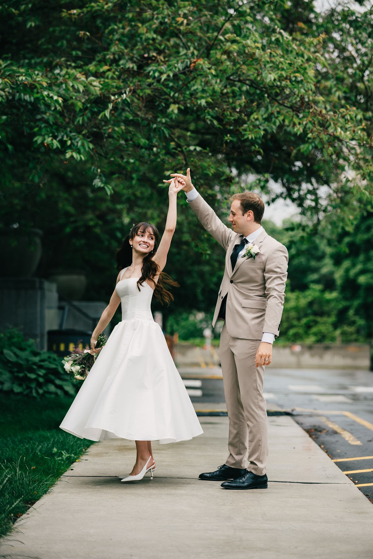 A couple dances playfully on a sidewalk surrounded by lush green trees and bushes.