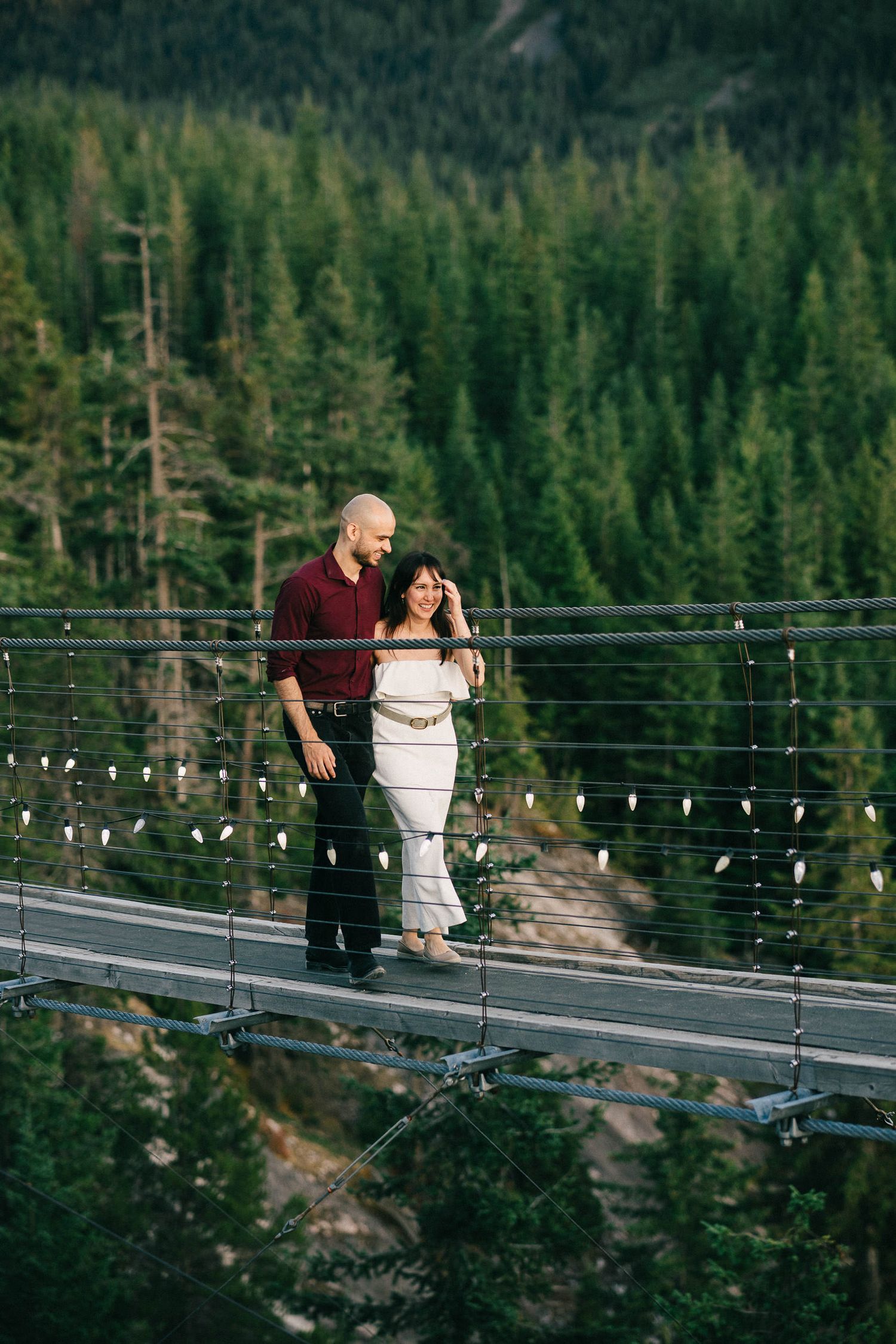 A couple stands together on a suspension bridge over a forest with string lights overhead.