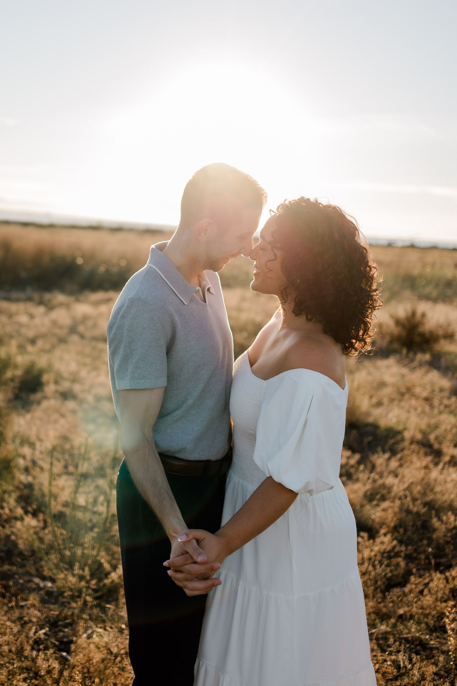 A romantic couple embraces in a sunlit field at golden hour as the sun sets behind them creating a warm glow.