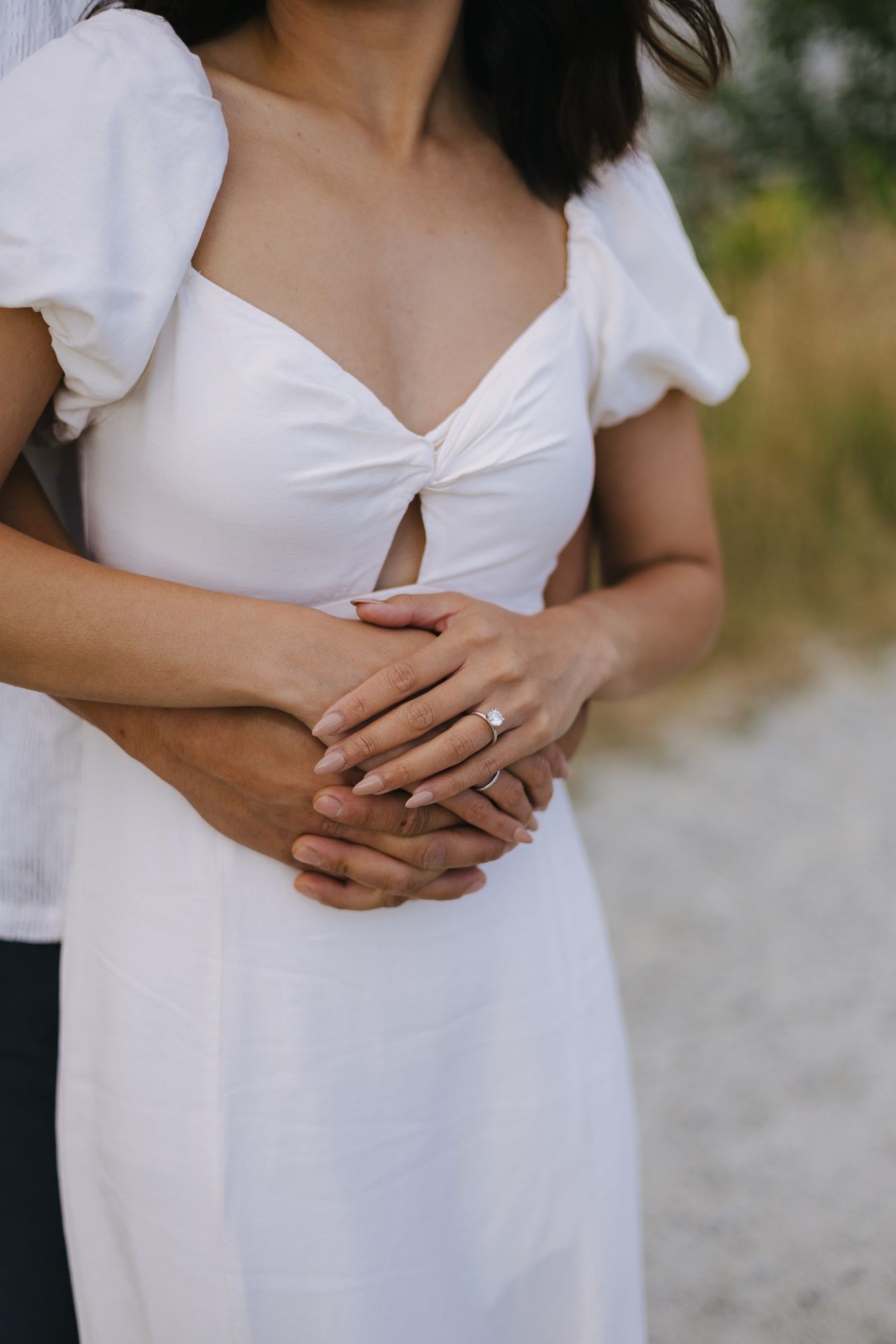 Close-up of a woman in a white dress with clasped hands wearing an engagement ring as her partner hugs her from behind.