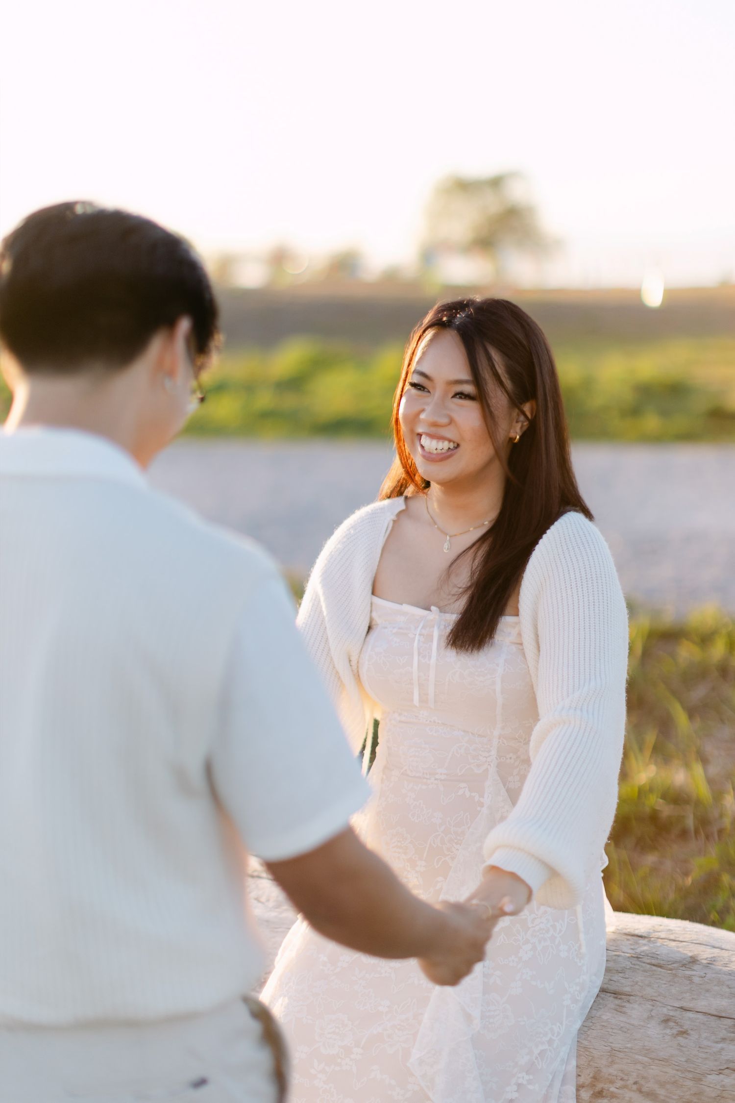 Smiling woman in a white lace dress and cardigan holds hands with her partner outdoors at golden hour sunset.