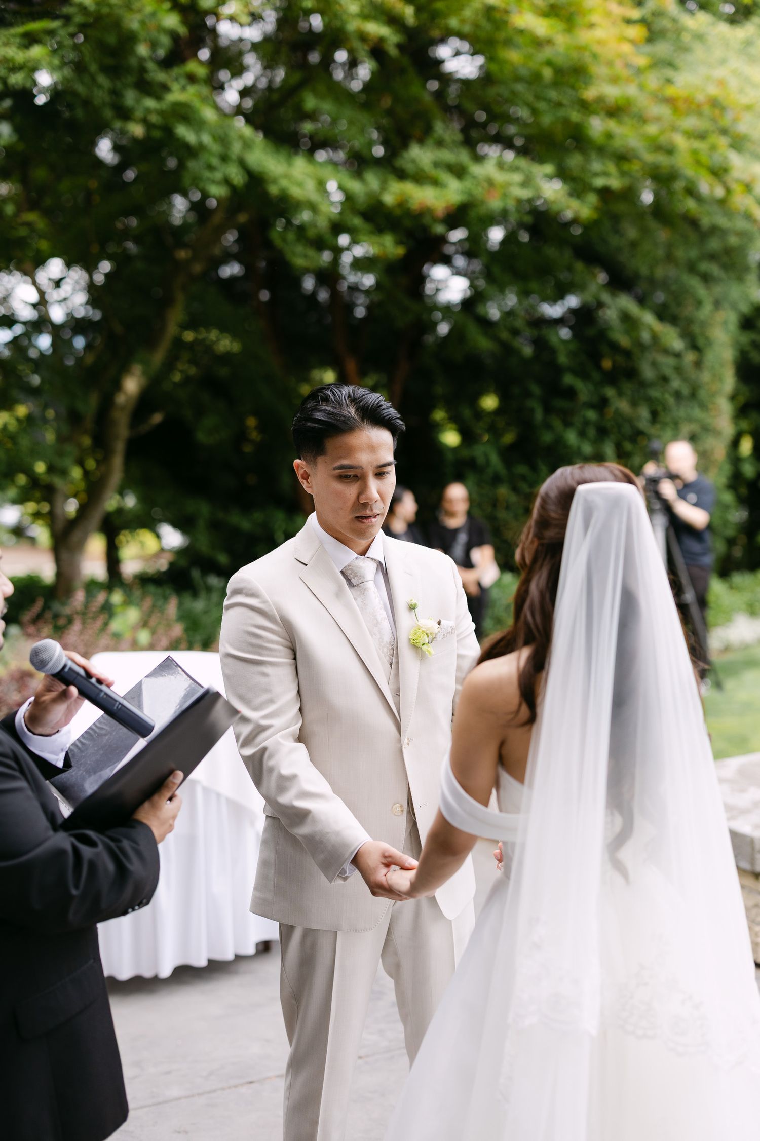 Groom in beige suit holds bride's hands during outdoor wedding ceremony while officiant reads vows.