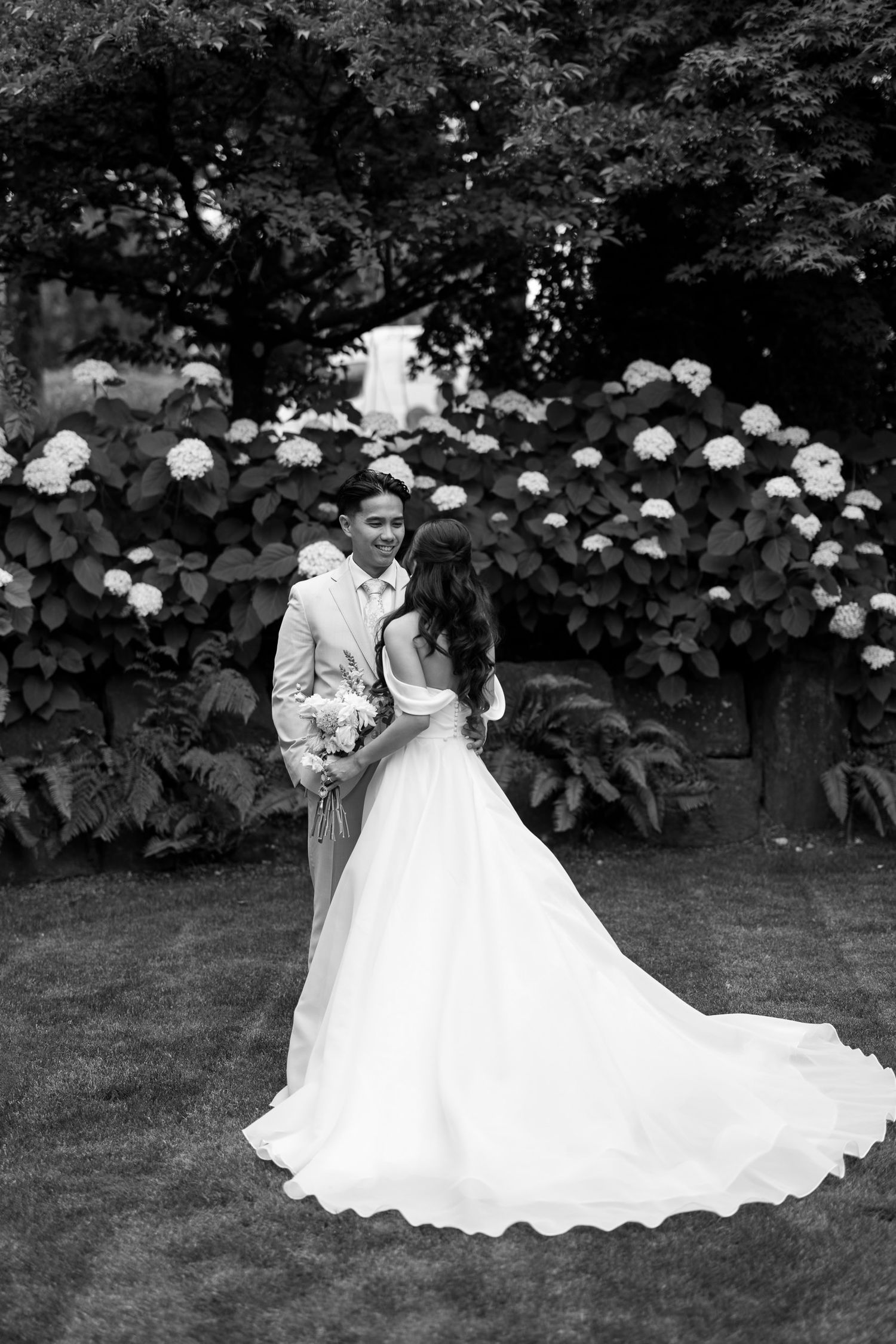 Black and white photo of bride in flowing gown and groom embracing before hydrangea bushes outdoors.