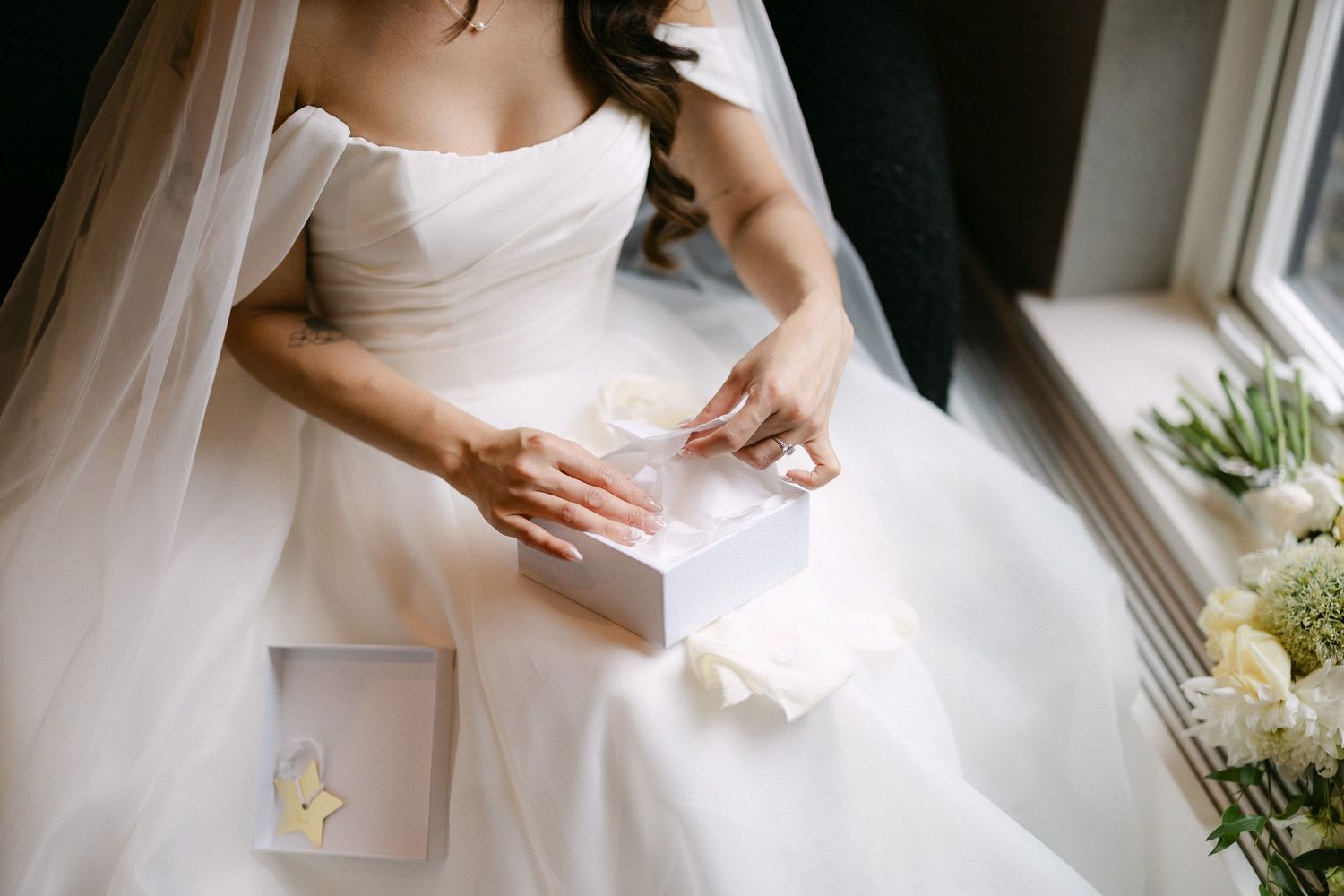 Bride in white strapless wedding gown opening a small gift box while seated near a window on her wedding day.
