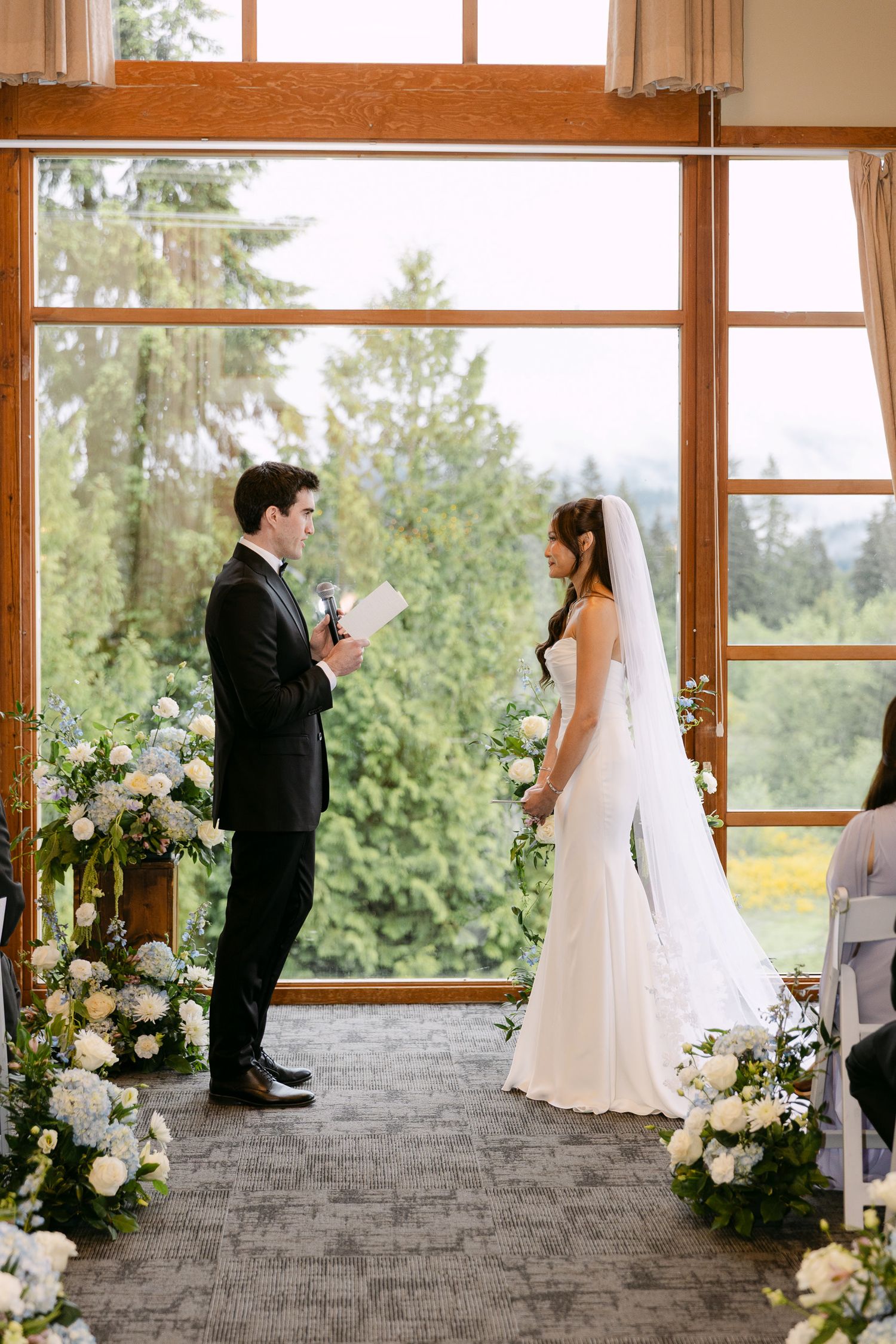 Bride and groom exchange vows at elegant indoor wedding ceremony with floral arrangements and large windows overlooking lush greenery.