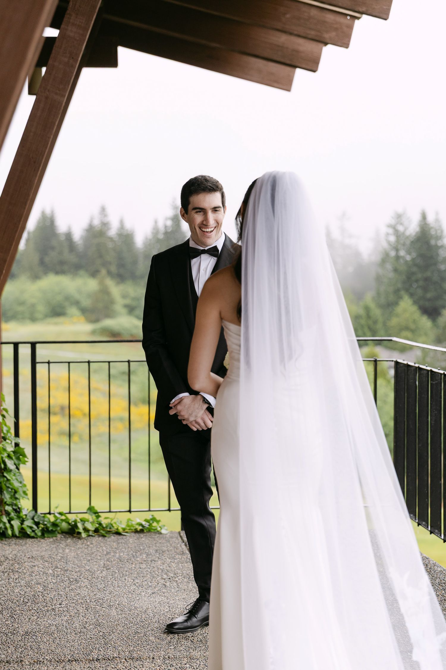 Groom in black tuxedo smiles at bride in white gown and veil on outdoor venue balcony with lush green trees backdrop.