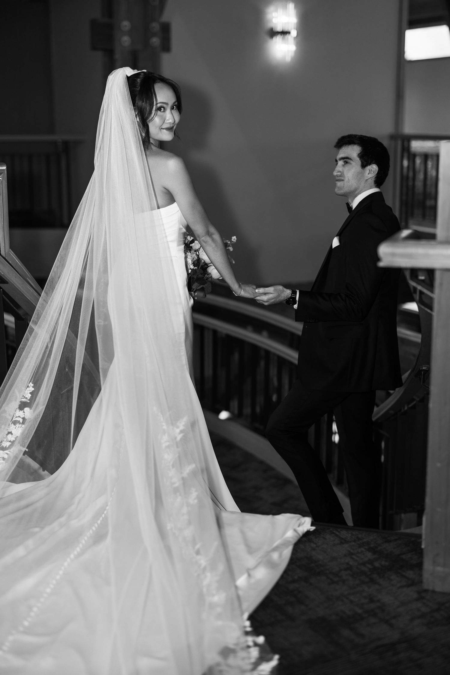 Black and white photo of a bride in a long veil and groom in a tuxedo holding hands at a church altar.
