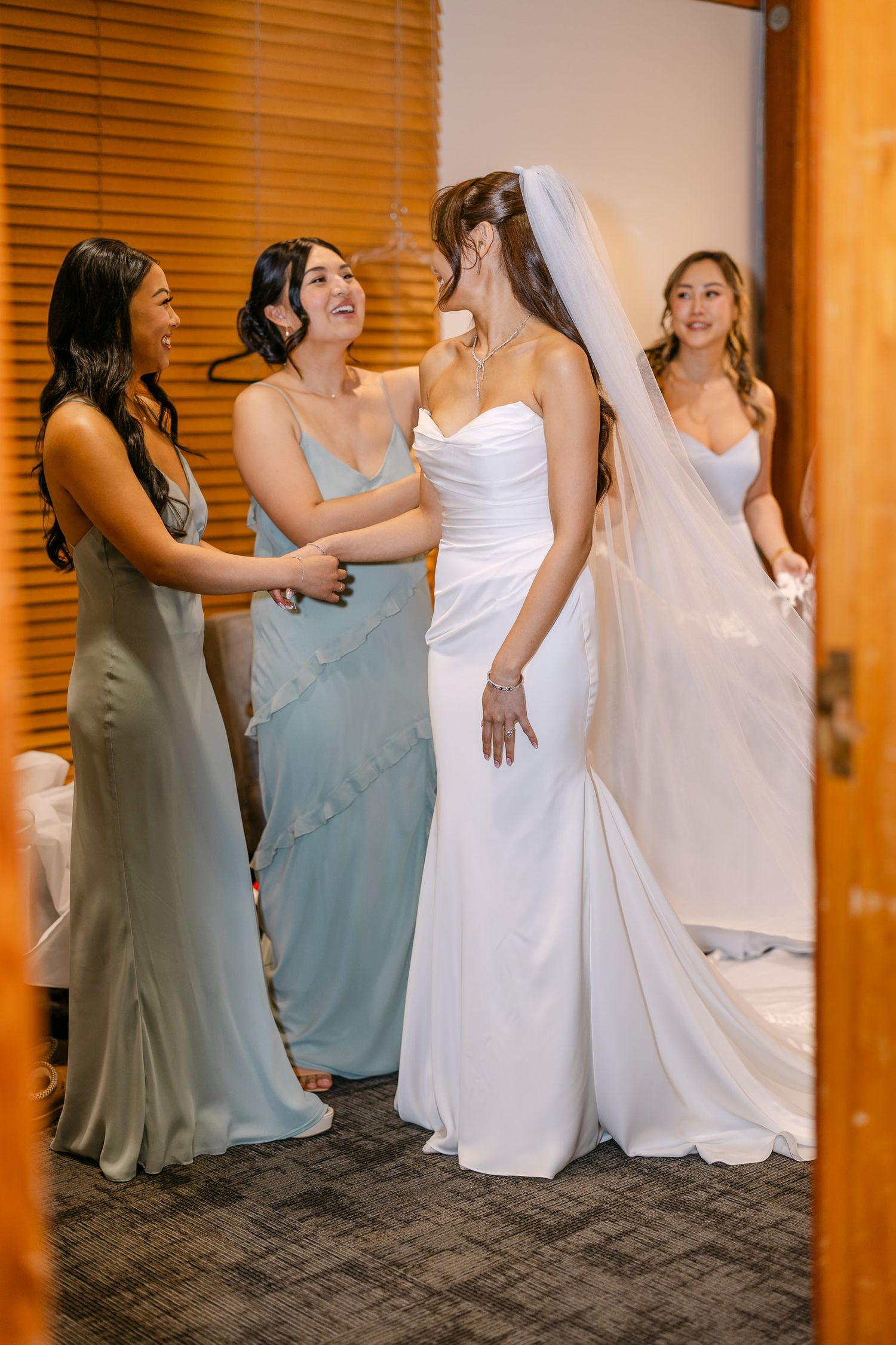 Bride in white gown and veil surrounded by bridesmaids in sage green dresses during wedding preparation.
