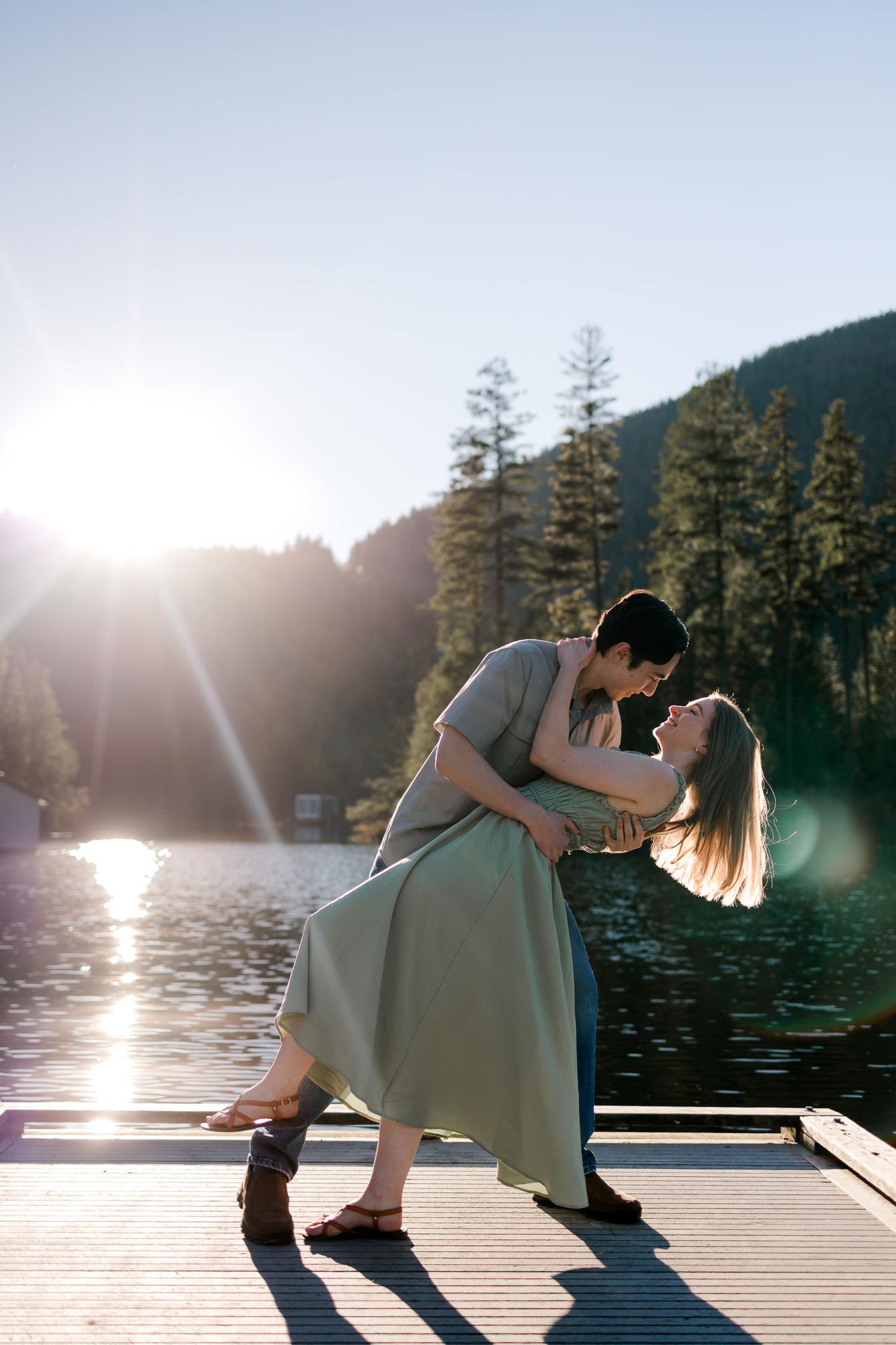 A romantic couple dances on a lakeside dock at sunset with tall pine trees and mountains in the background.