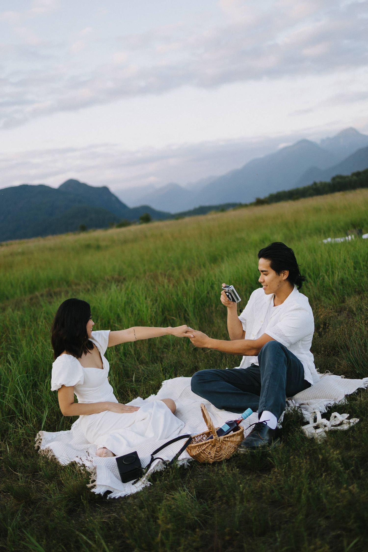 Two people relax together in a grassy meadow with mountains visible in the background on a cloudy day.