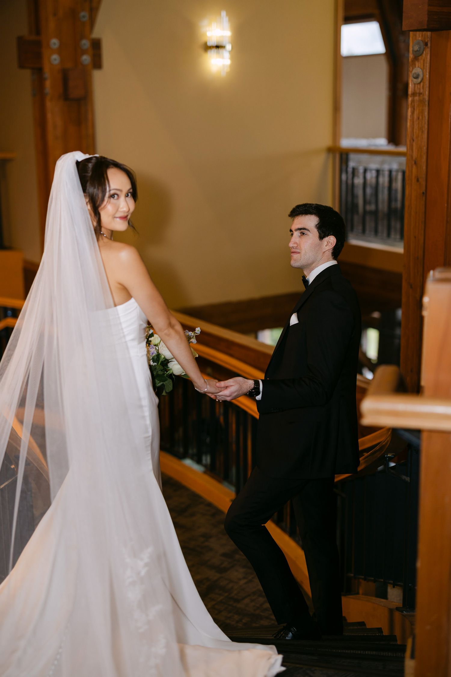 Bride in cathedral veil and groom in tuxedo hold hands on elegant wooden staircase.