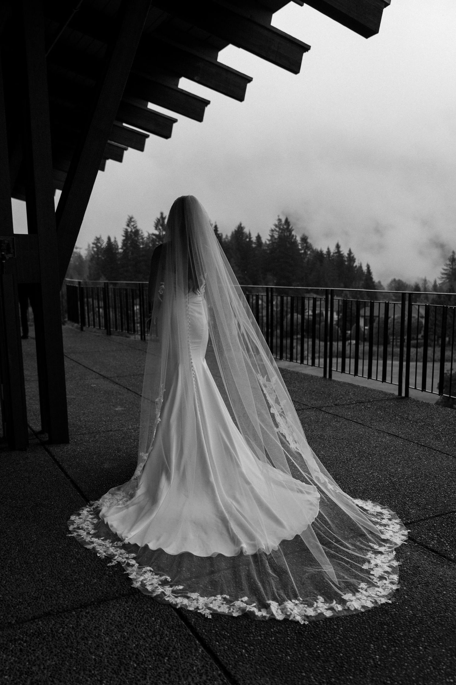 Black and white photo of a bride in a flowing gown with lace-trimmed cathedral veil, standing on an outdoor terrace.