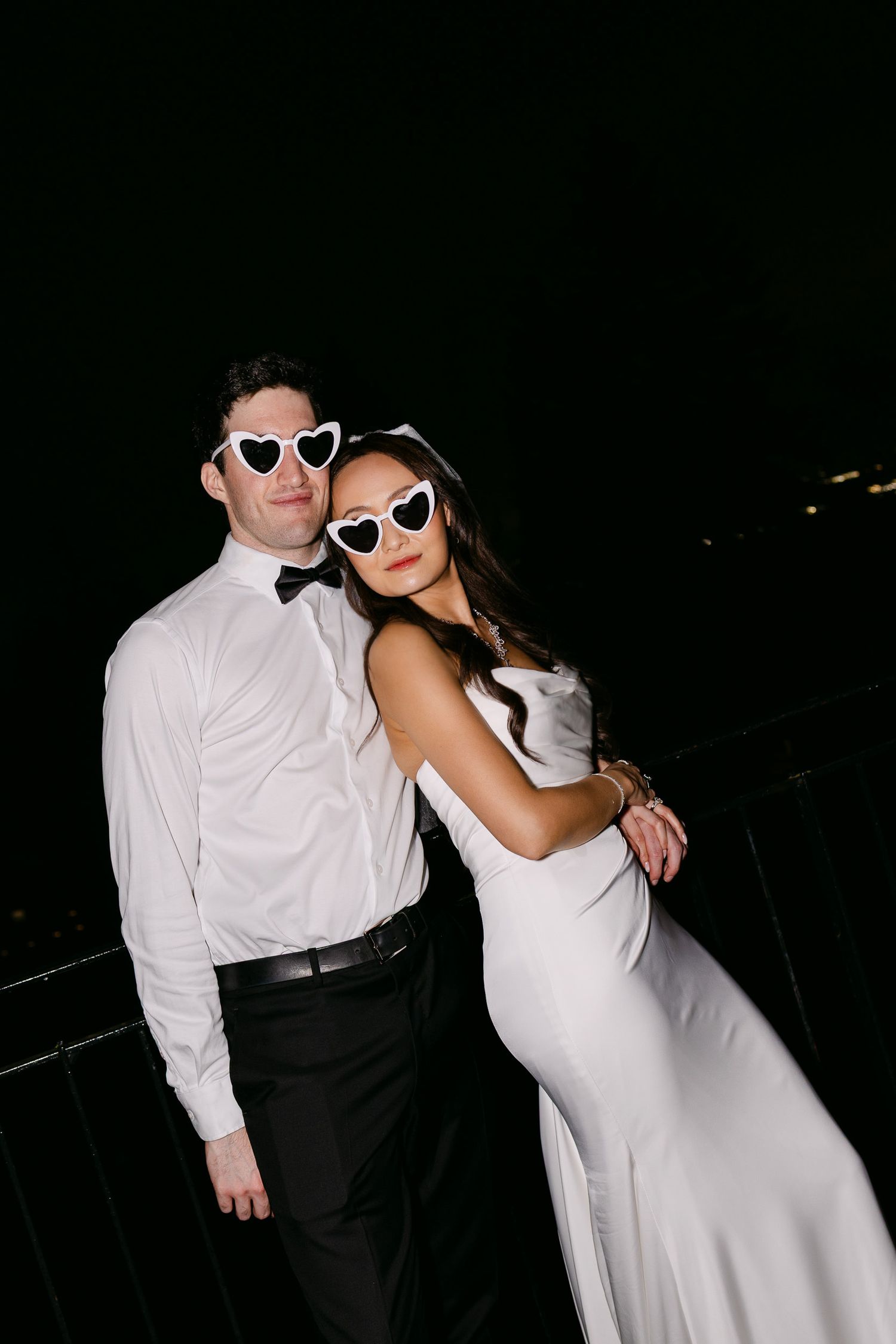 Couple in white heart-shaped sunglasses posing at night, man in tuxedo shirt and woman in elegant white wedding gown.