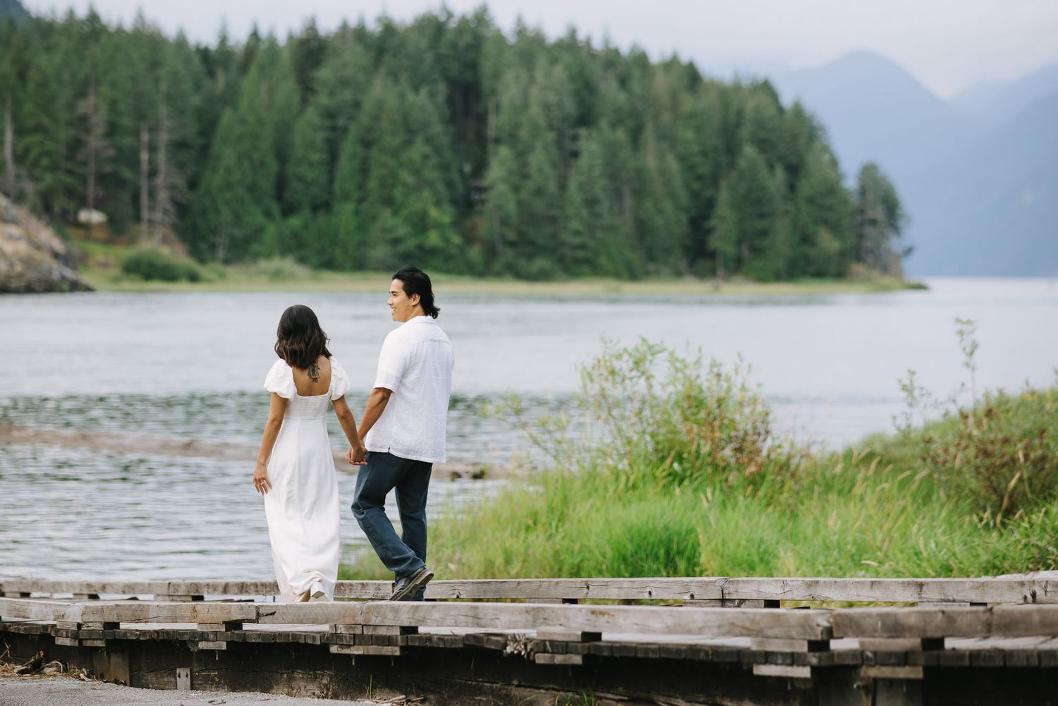Couple walks hand in hand along a wooden boardwalk beside a lake with a forested mountain backdrop.