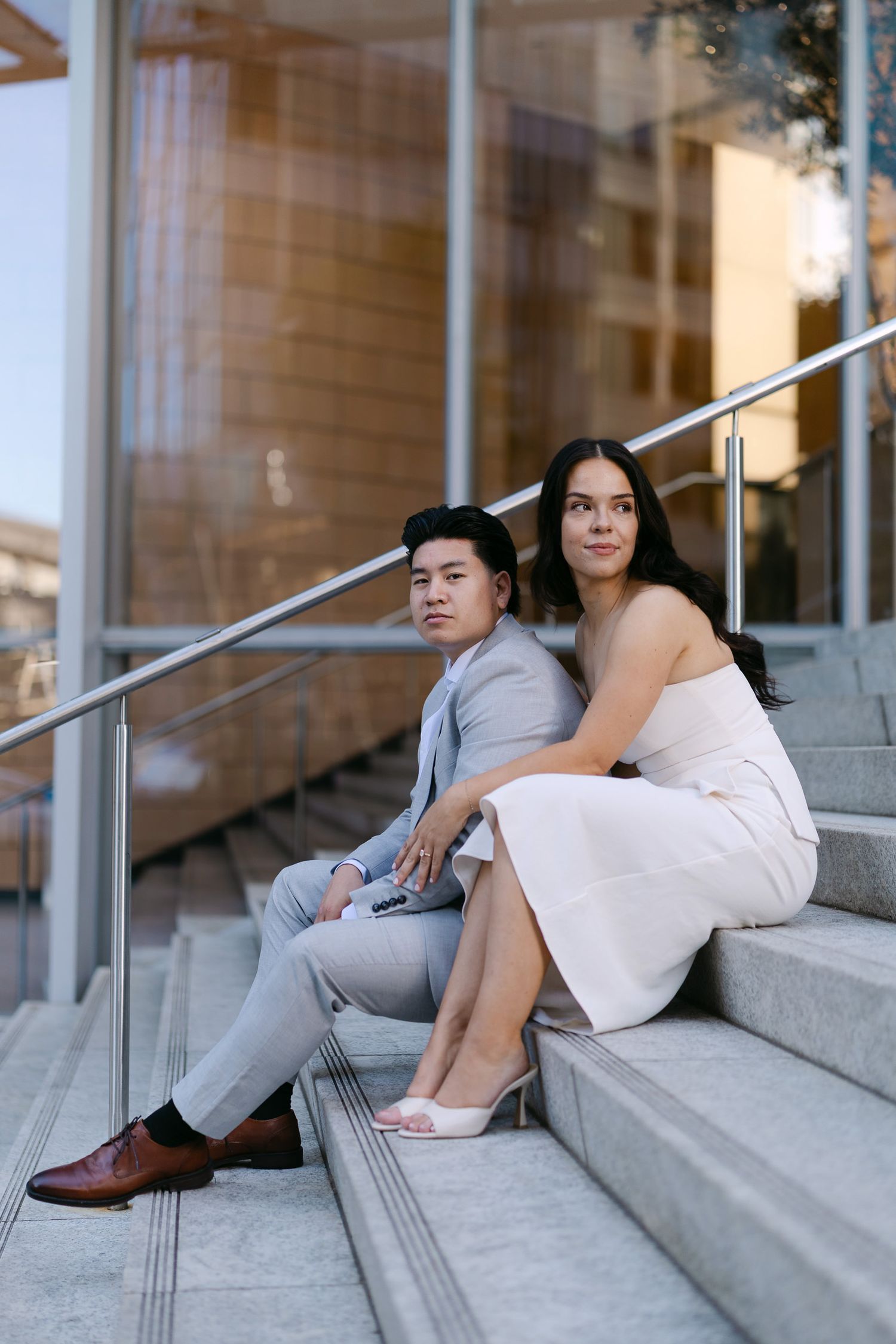 A couple sits close together on outdoor stone steps near a glass building, the man in a gray suit and the woman in a white dress.