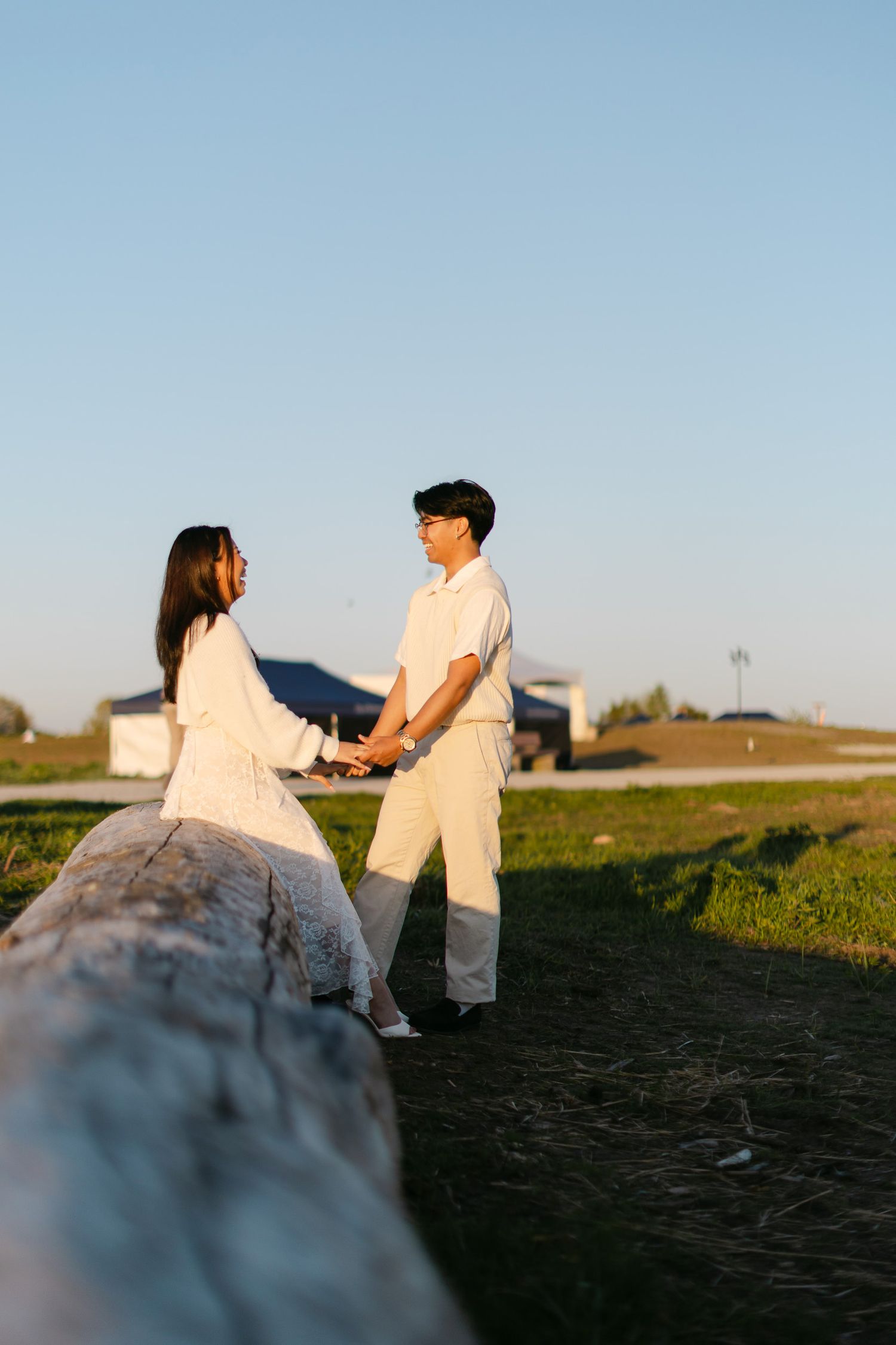 Couple in white outfits holding hands at golden hour in a grassy field beside a weathered log under a clear blue sky.