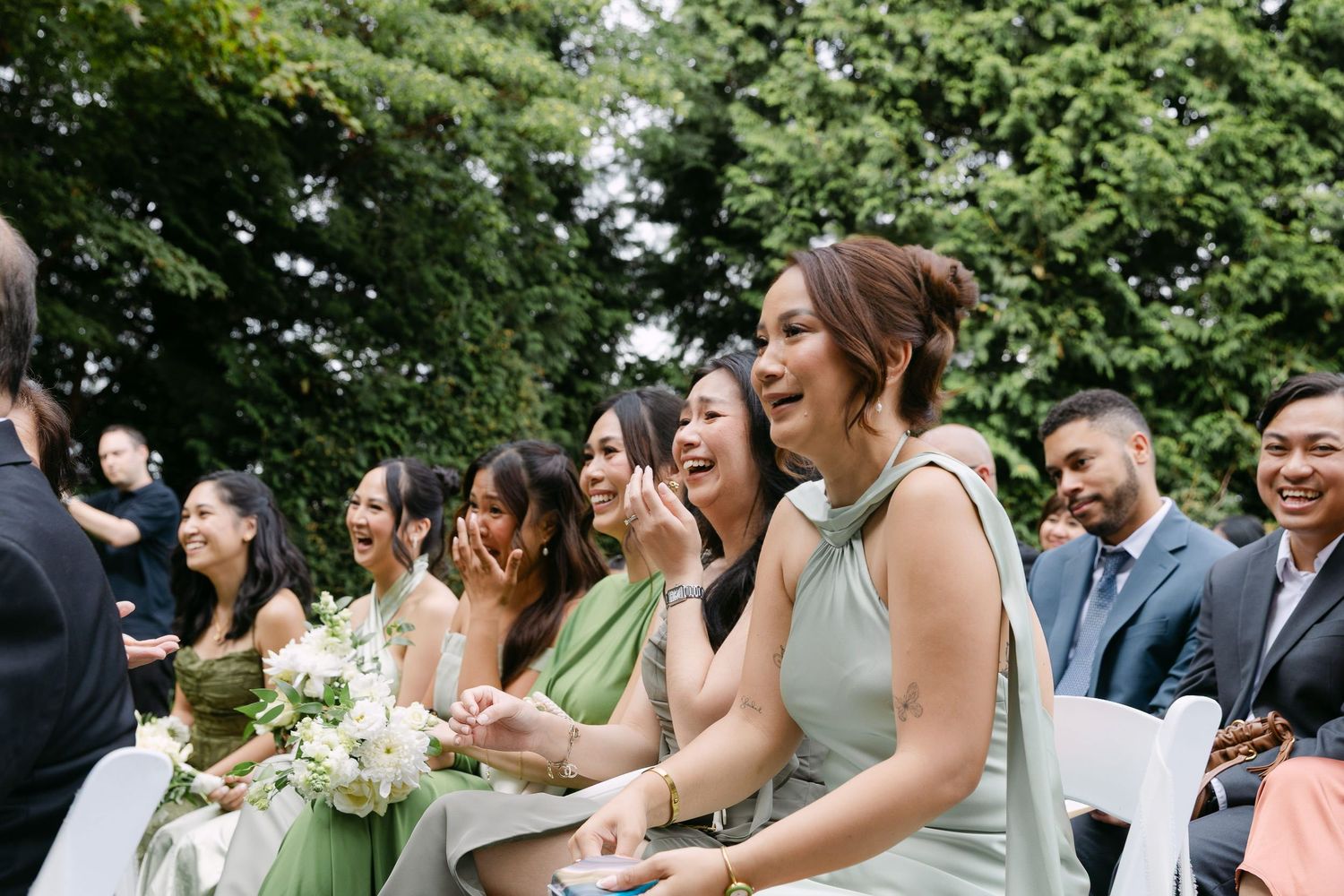 Bridesmaids in sage green dresses laughing joyfully during an outdoor wedding ceremony.