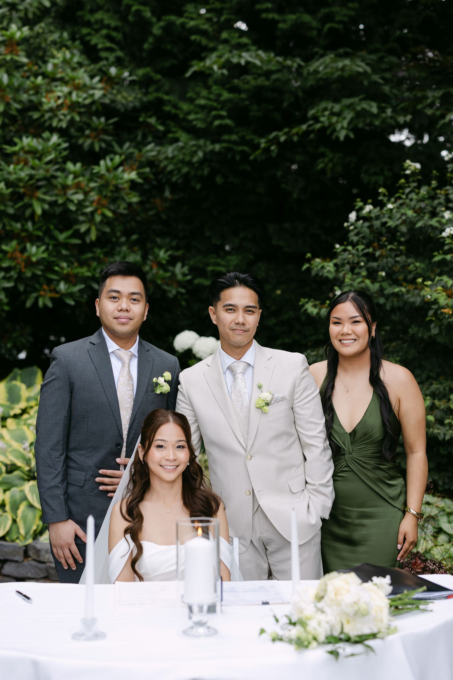 A bride and groom pose with two guests at an outdoor wedding reception table surrounded by lush greenery.