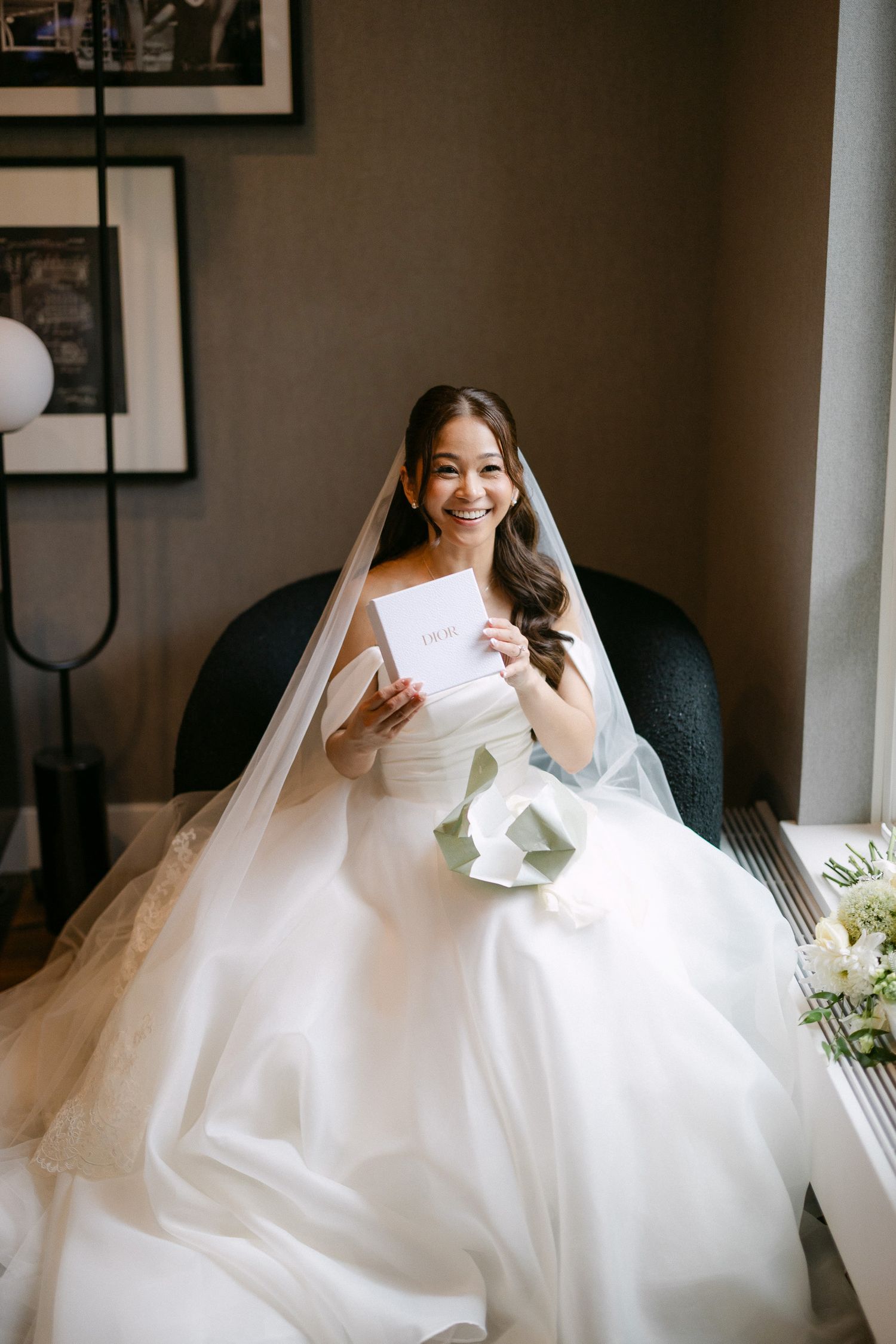 Smiling bride in white ball gown and veil reads a letter while seated, holding a bouquet on her wedding day.