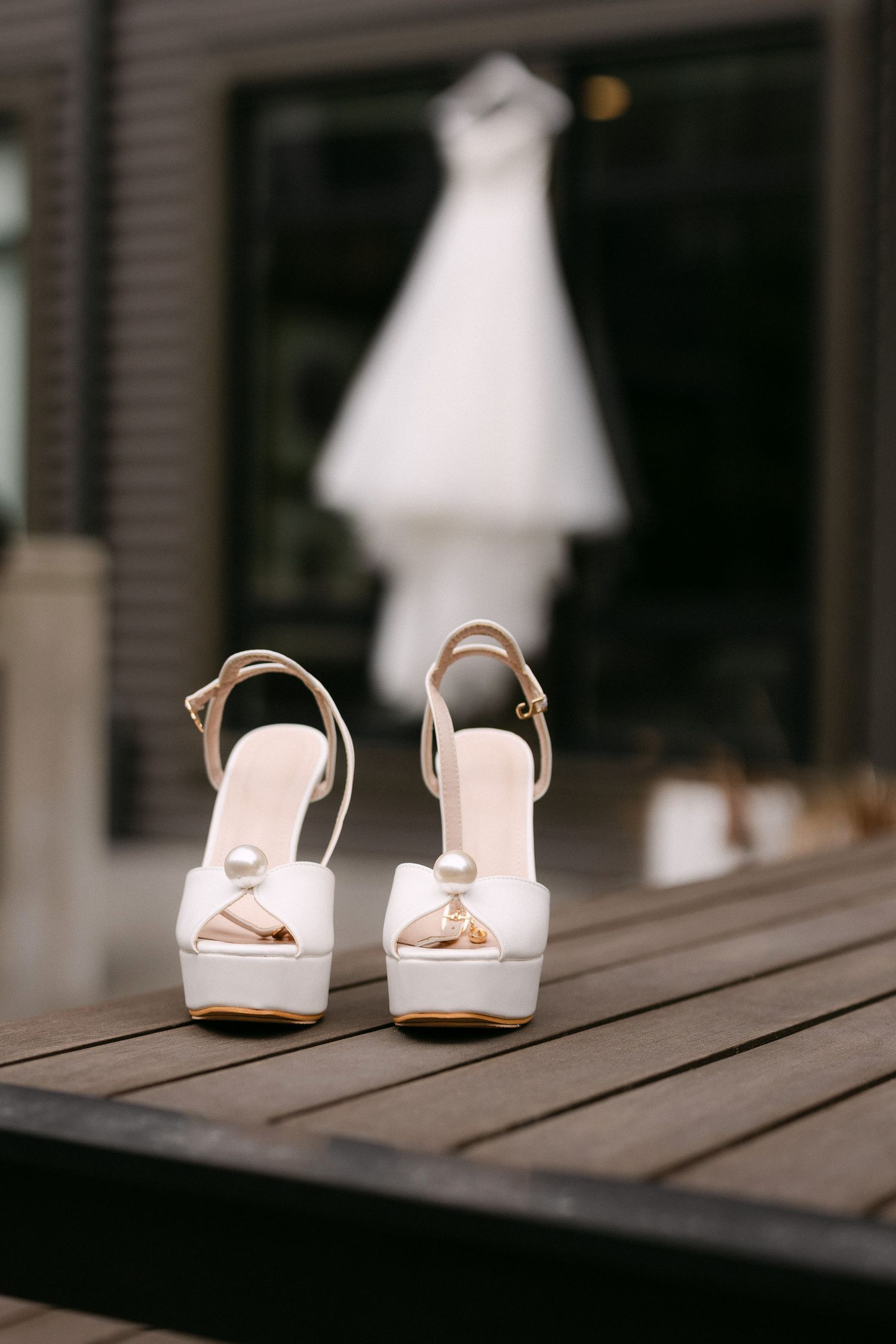 White bridal platform heels with pearl accents on a wooden surface, with a wedding dress hanging blurred in the background.