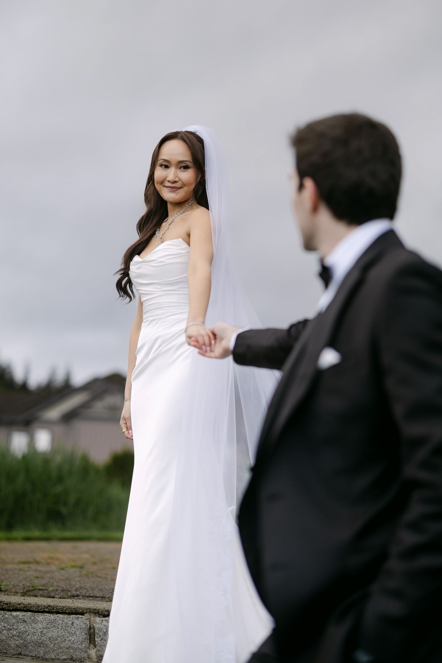 Bride in white strapless gown and veil holds grooms hand outdoors on cloudy day.