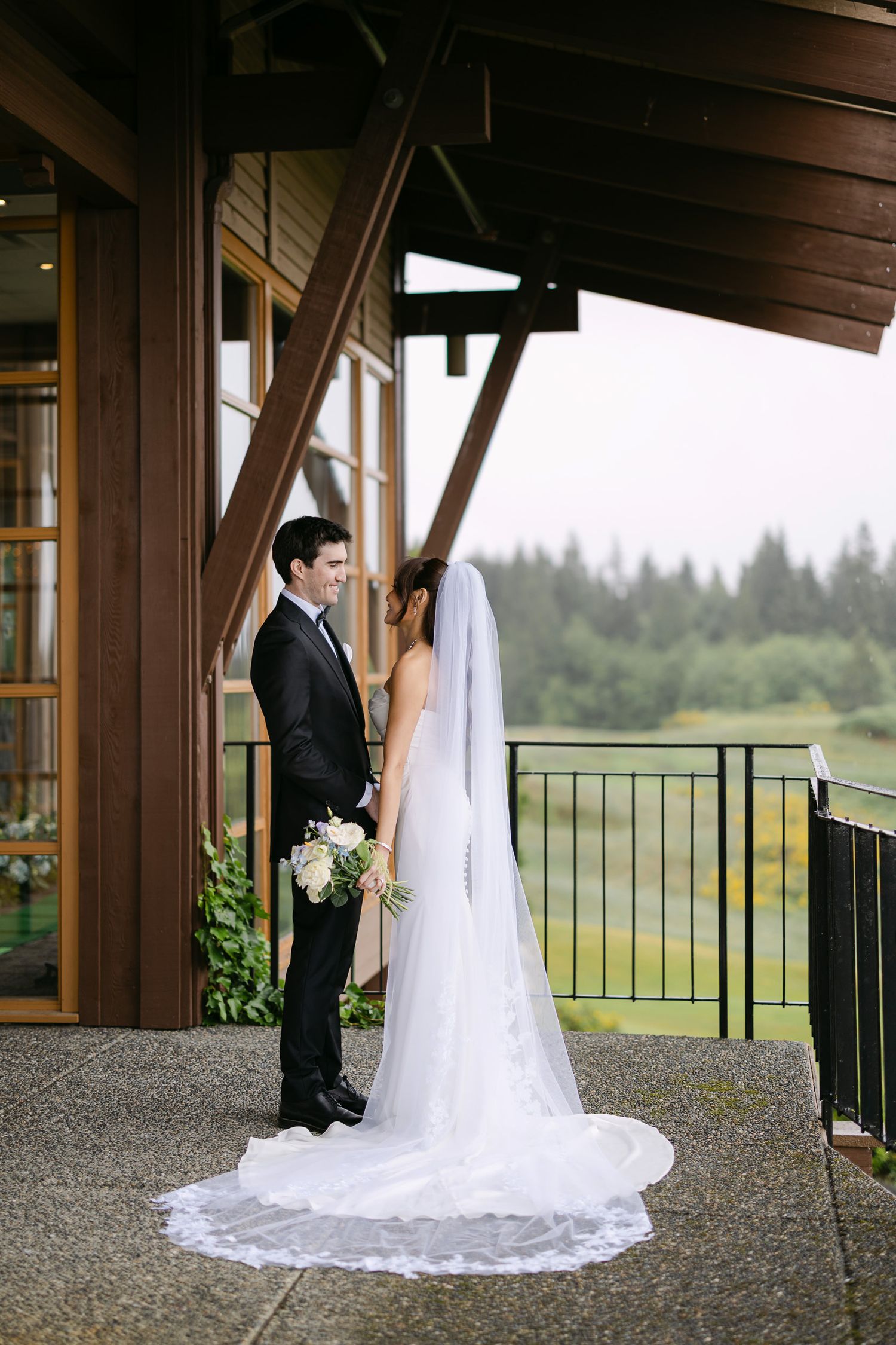 Bride and groom share a romantic moment on a venue balcony, her lace cathedral veil trailing elegantly.