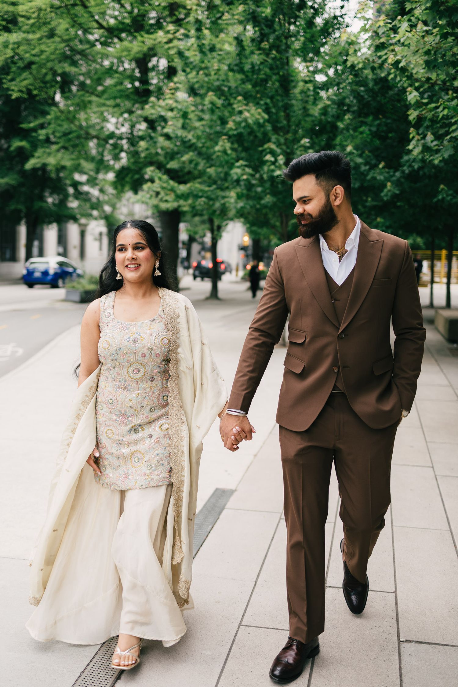 A couple in coordinated formal wear walks hand in hand along a tree-lined urban pathway.