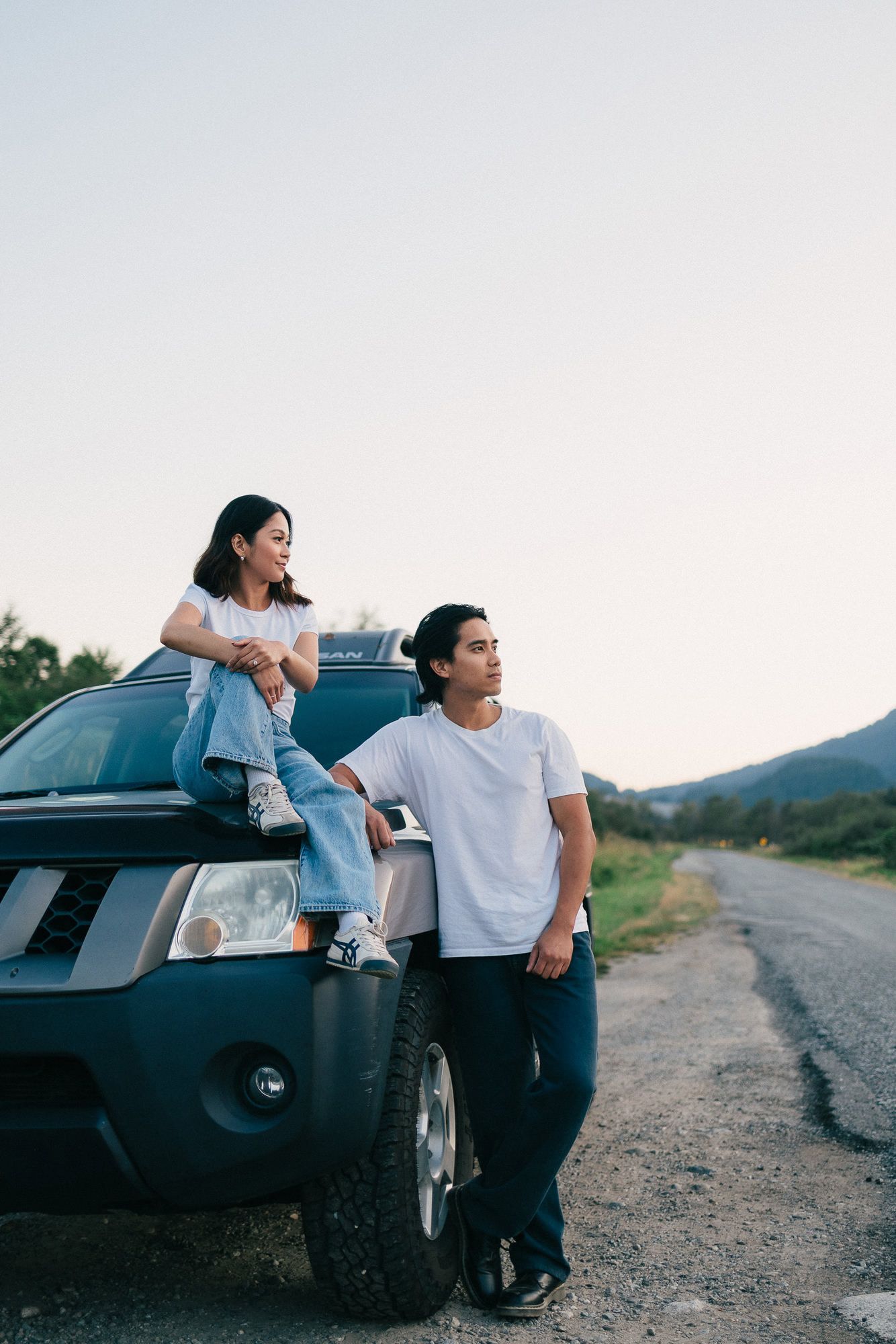 A couple poses casually by a dark SUV on a country road at dusk with mountains in the distance.