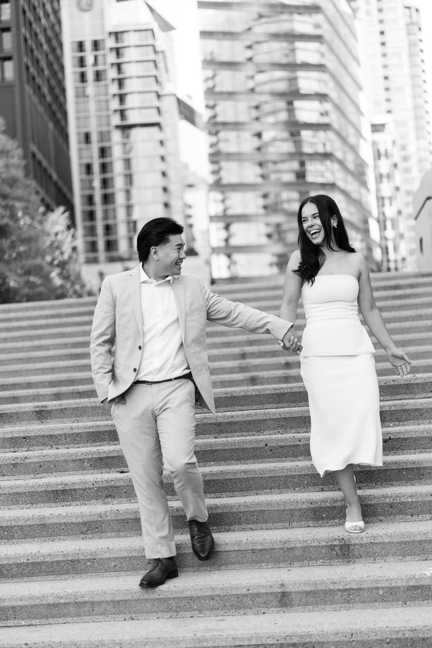 Smiling couple holding hands and walking down concrete steps in a downtown city setting, in black and white.