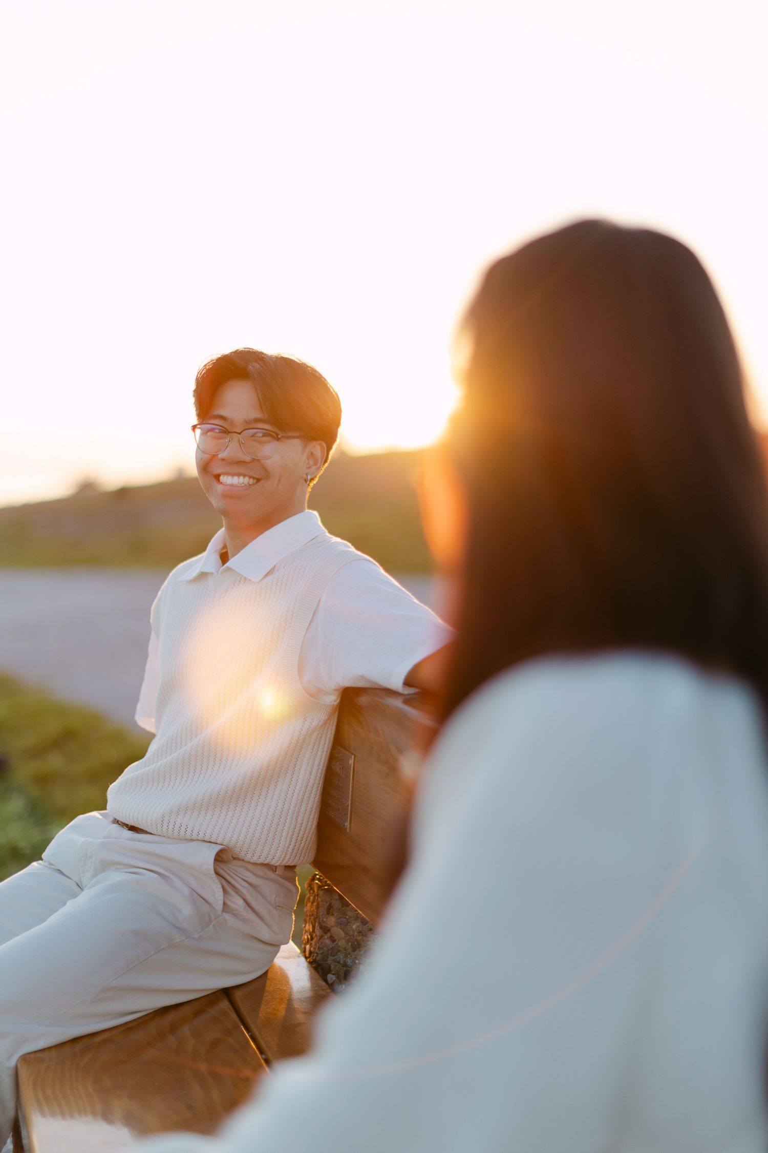 Smiling man in glasses and a cream polo sits on a wooden bench at golden hour as a woman faces him in the foreground.