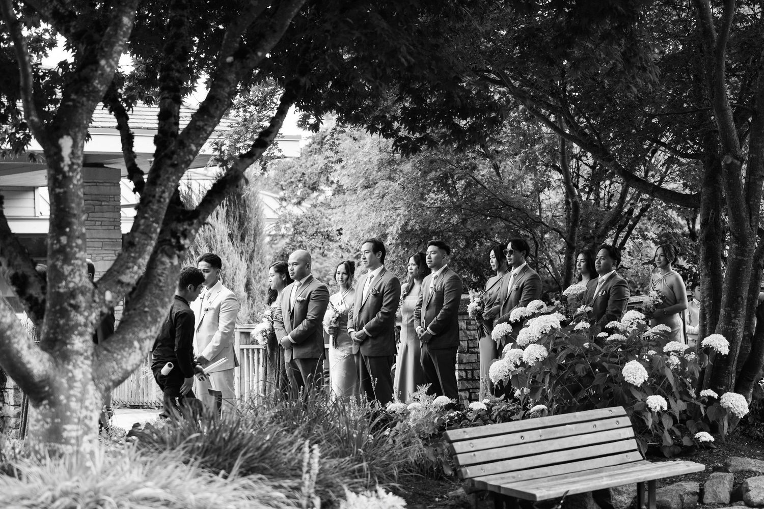 Black and white photo of a group of people standing outdoors among trees and flowers at a garden gathering.