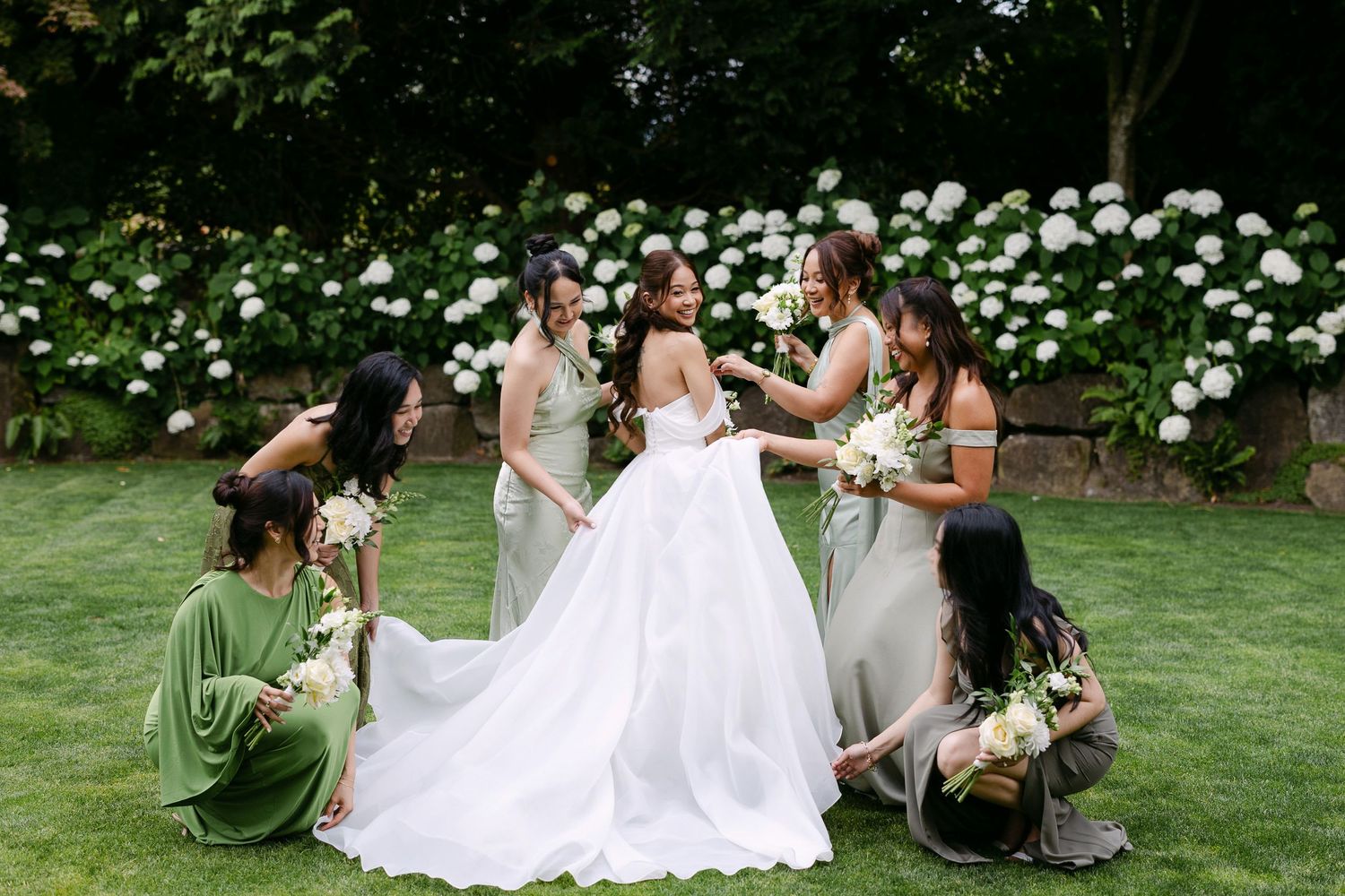 Bride in white gown surrounded by bridesmaids in green and gray dresses holding bouquets on lush garden lawn.