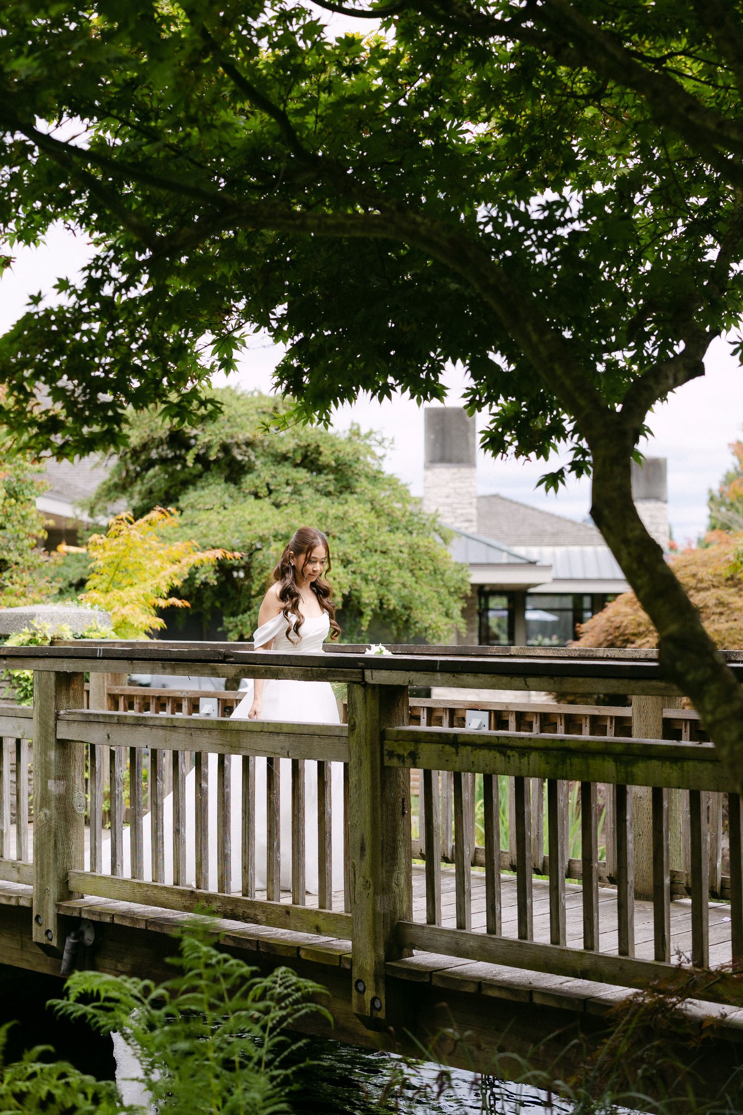 Bride in white dress standing on a wooden garden bridge surrounded by lush green trees and foliage.