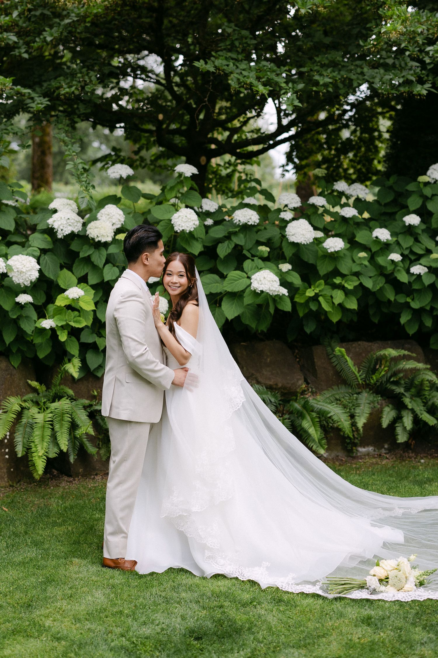 Couple in wedding attire embracing in a lush garden with white hydrangeas in bloom.